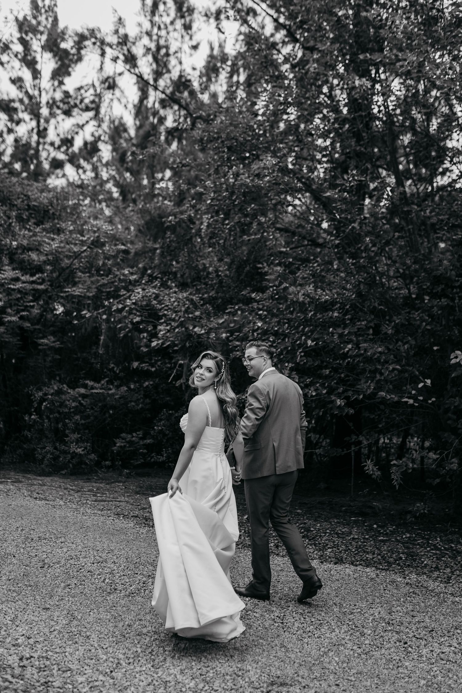 A black and white photo of newlyweds walking together on a gravel path surrounded by trees.