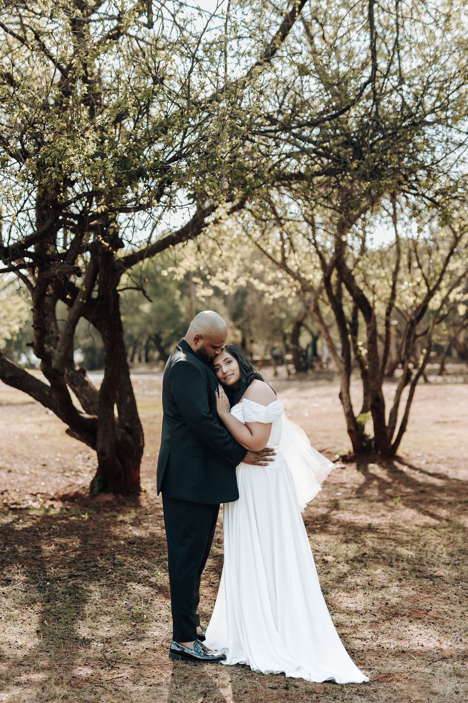 Intimate wedding moment captured under flowering trees in a rustic outdoor setting.