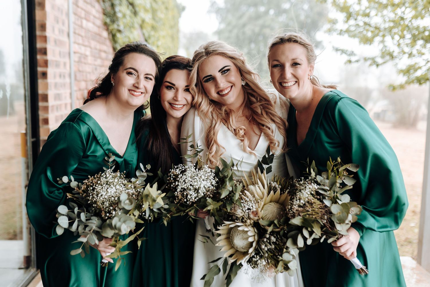 Happy bride and bridesmaids in white and emerald dresses holding matching floral bouquets.