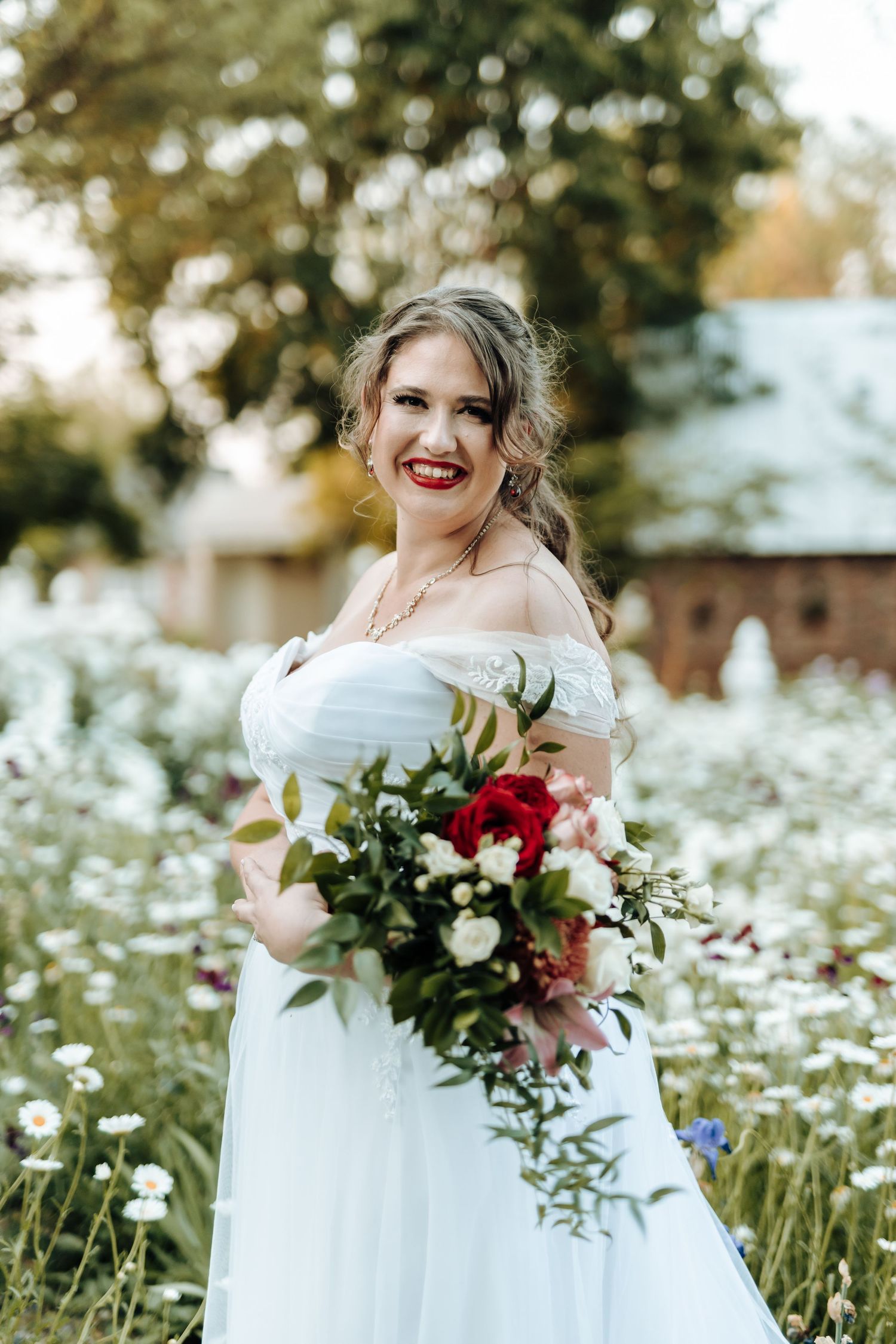 Off-shoulder wedding dress with cascading red and white bouquet in field of white wildflowers.