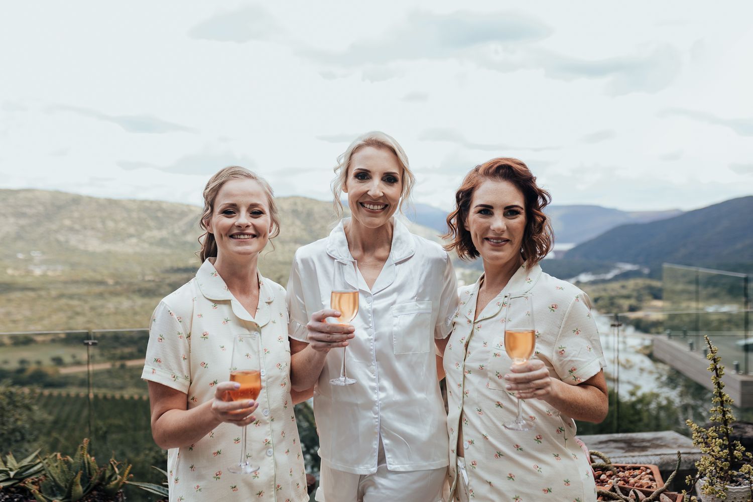 Three people in white attire toasting with champagne on mountain terrace.