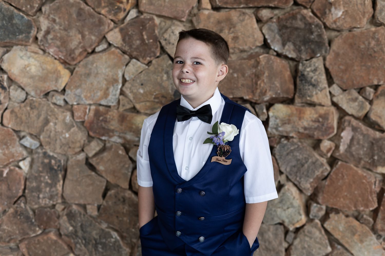 Young attendant in navy vest and bowtie poses against stone wall with boutonniere.