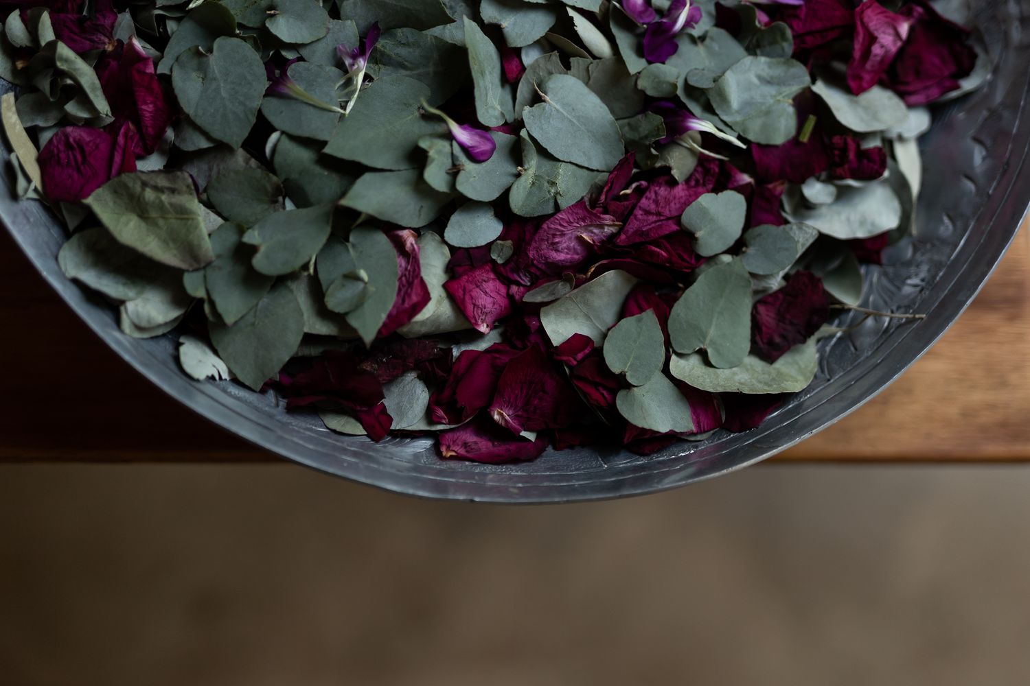 A bowl filled with green eucalyptus leaves and deep purple rose petals viewed from above.