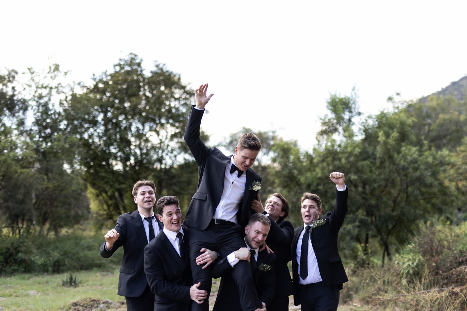 Energetic wedding party photo with groomsmen lifting and celebrating outdoors.
