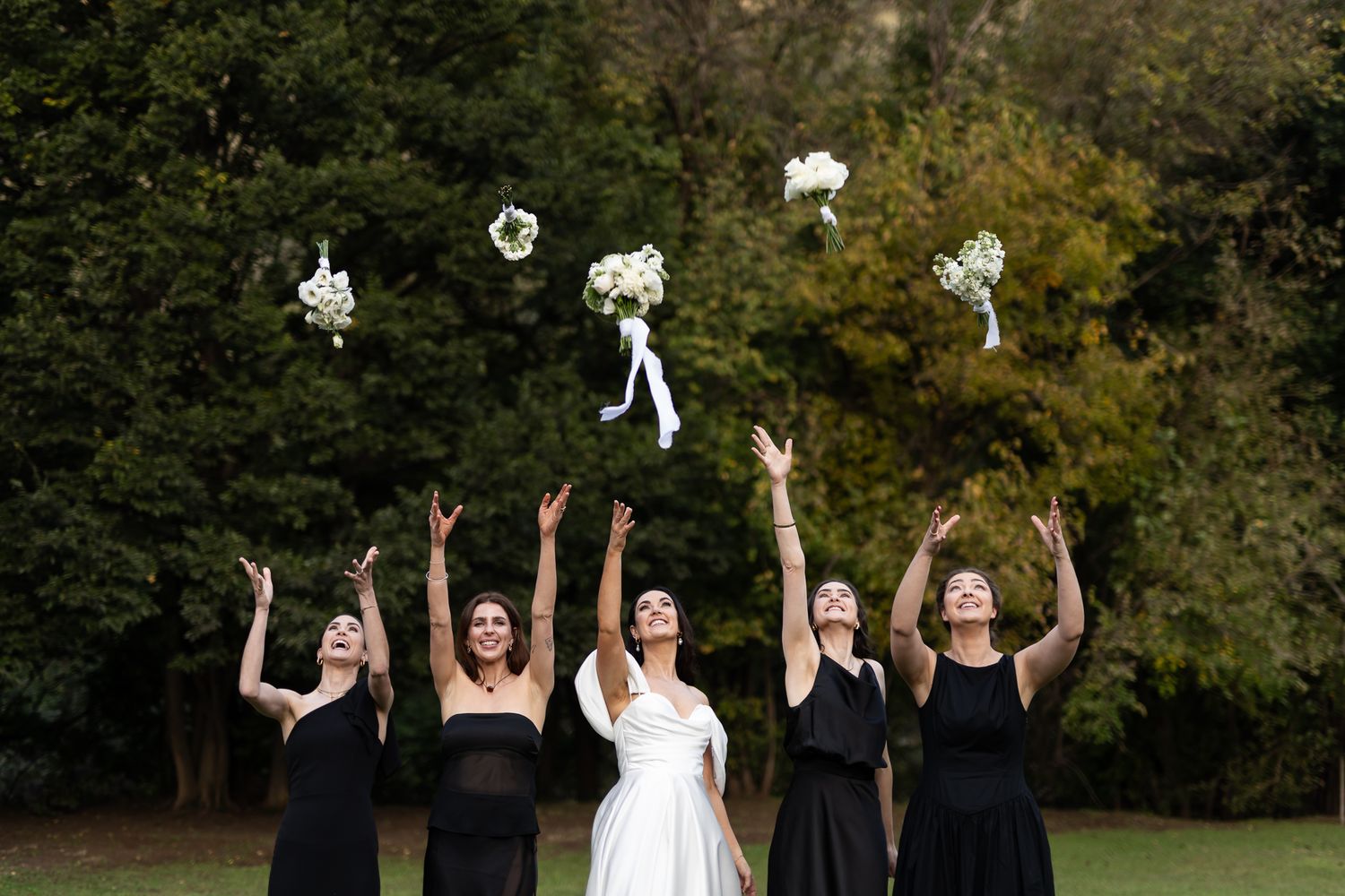 Wedding party in black dresses tossing white bouquets in the air against autumn trees.