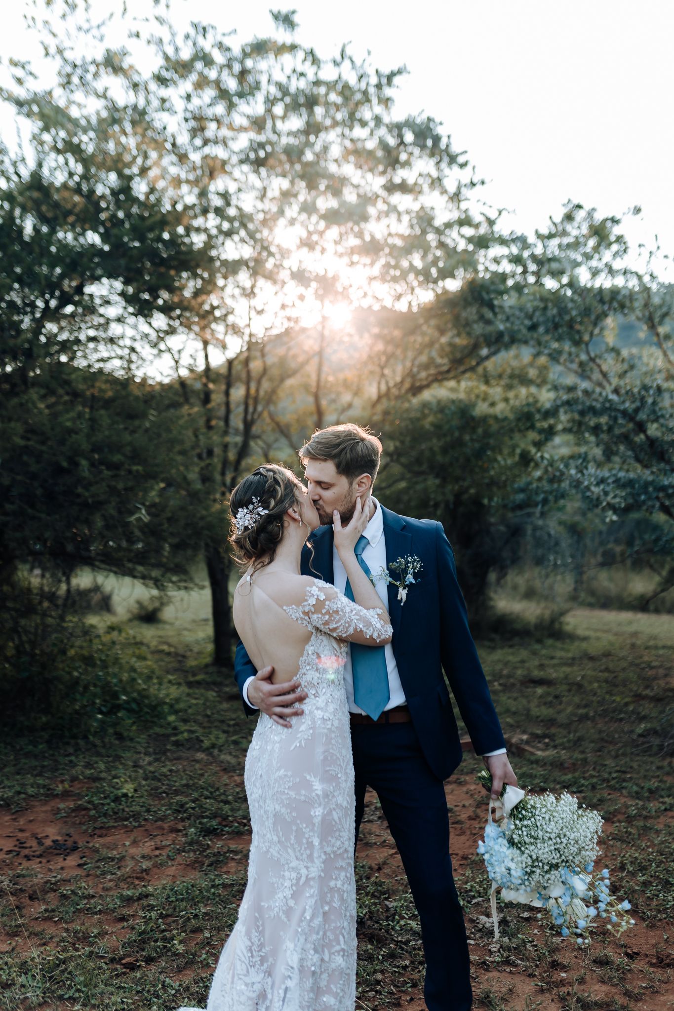 Romantic sunset wedding portrait with couple embracing among trees while holding white floral bouquet.