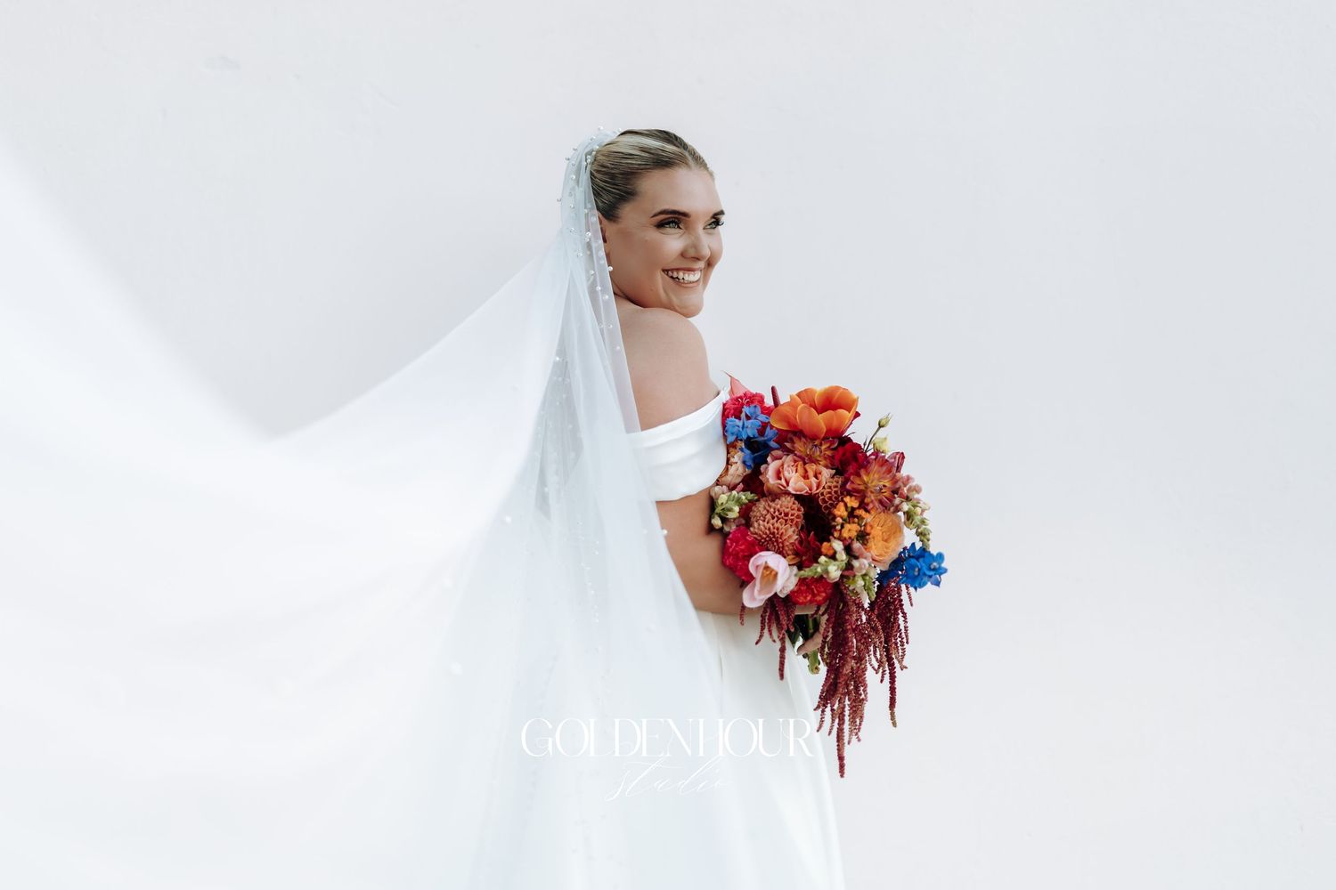 A bride in a white wedding dress holds a vibrant bouquet of orange, red and blue flowers against a light background.