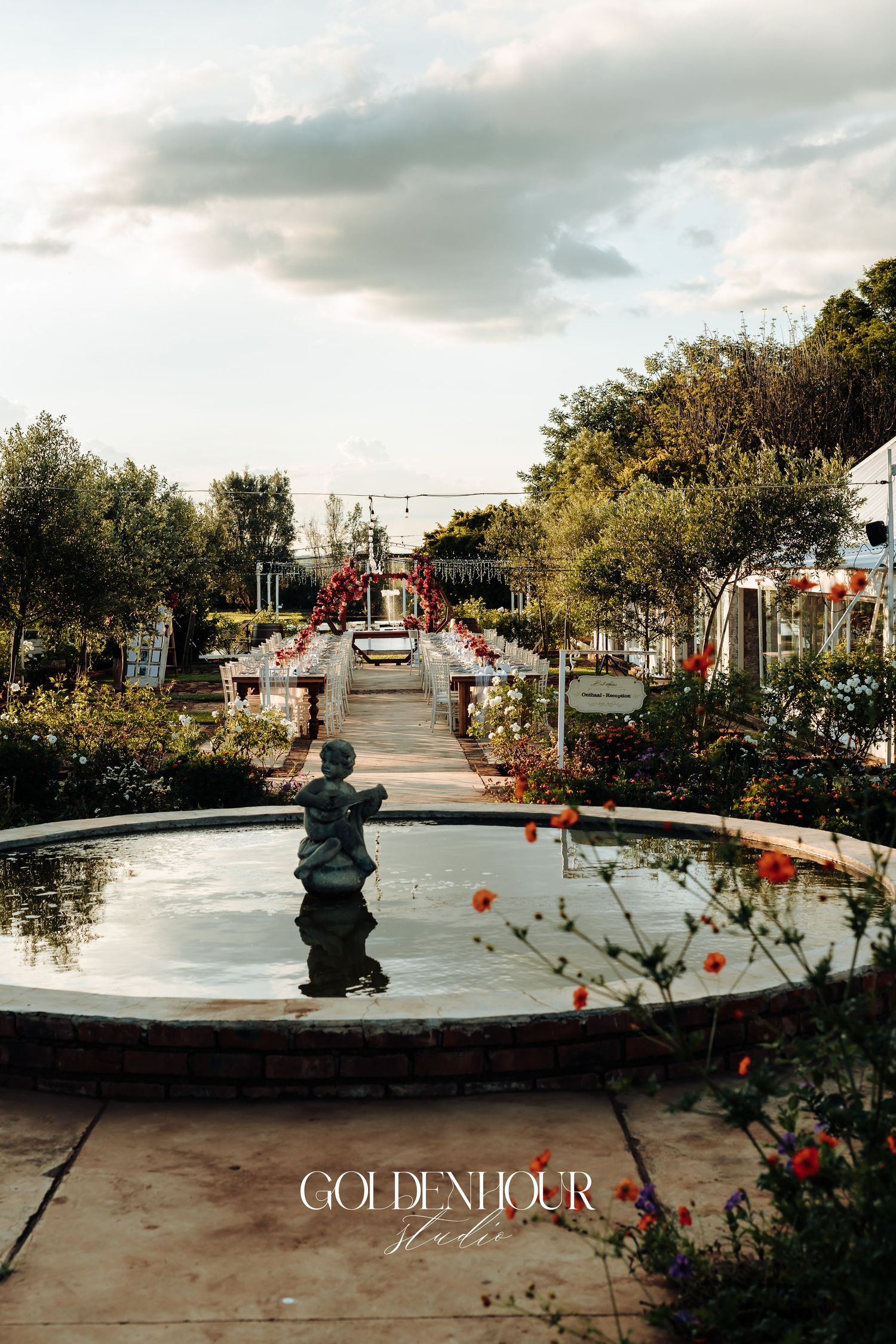 A serene garden walkway with a fountain and red flowers leads to a beachfront area under cloudy skies.