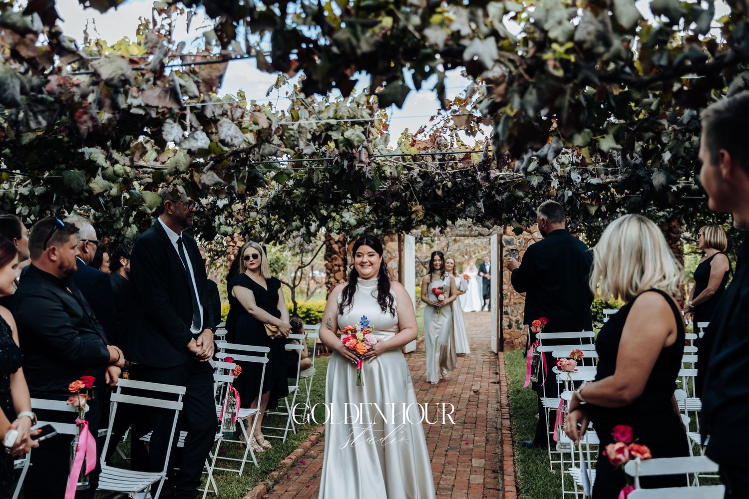 Beautiful outdoor garden wedding ceremony under floral archway with guests seated on black and white chairs.