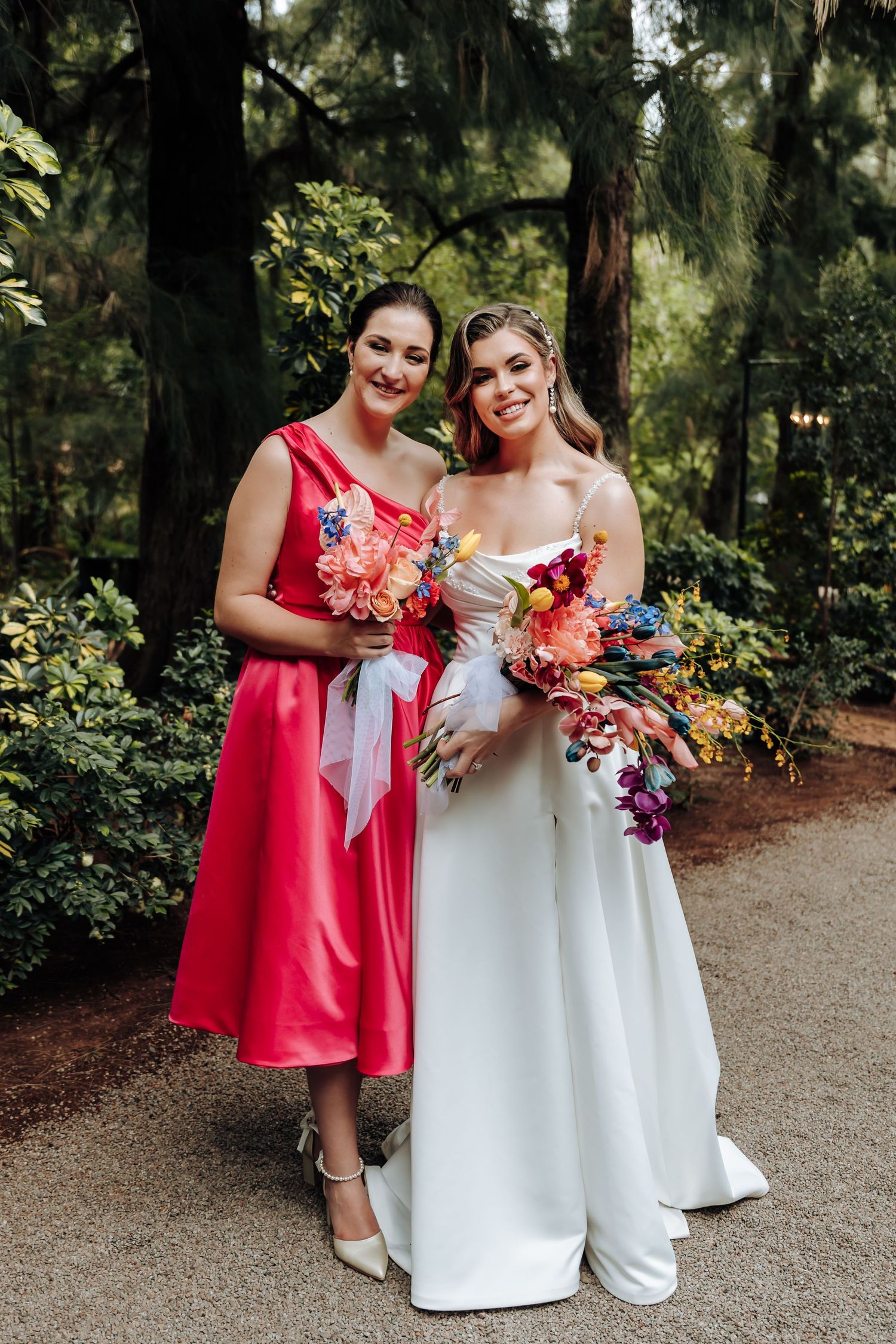 Two people in wedding attire pose together on a garden path while holding vibrant floral bouquets.