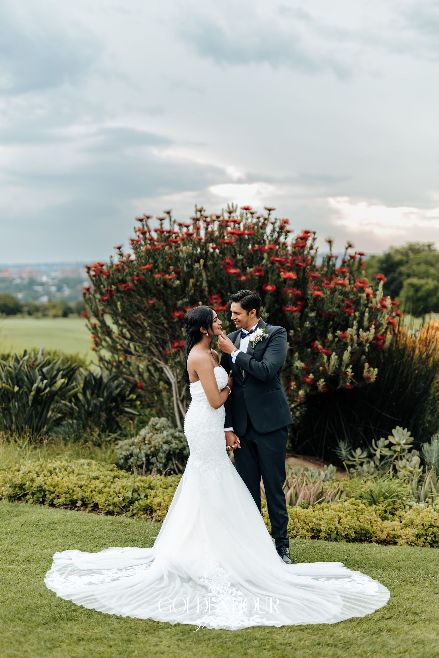Wedding couple embraces in garden setting with dramatic red flowering bush and cloudy sky.