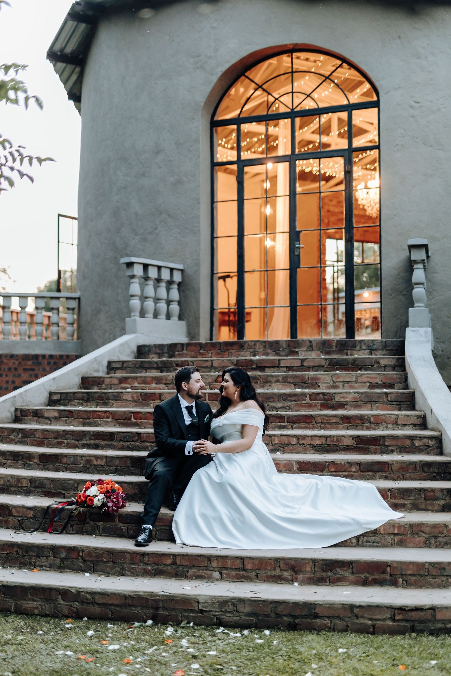 Wedding couple poses dramatically on stone steps with arched windows and warm interior lighting.