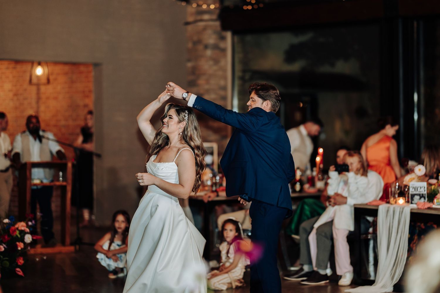 Wedding celebration on dance floor with guests dancing under ambient lighting in rustic venue.