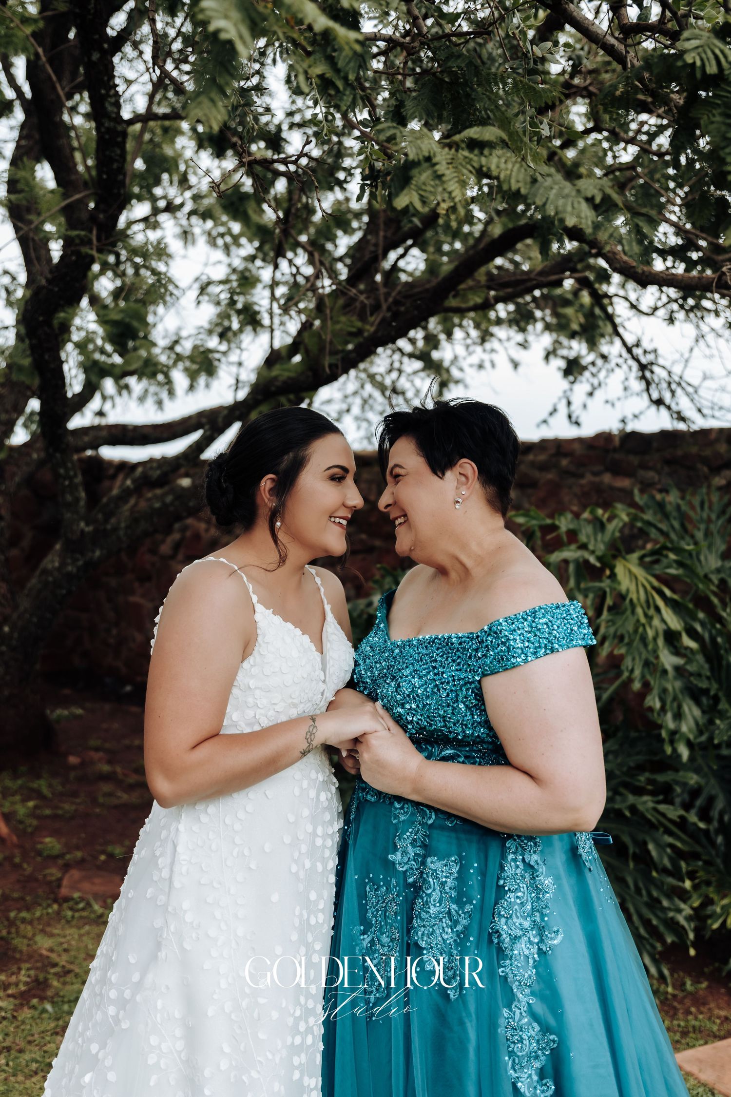 Two brides share intimate moment in white lace and turquoise sequined gowns under tree canopy.