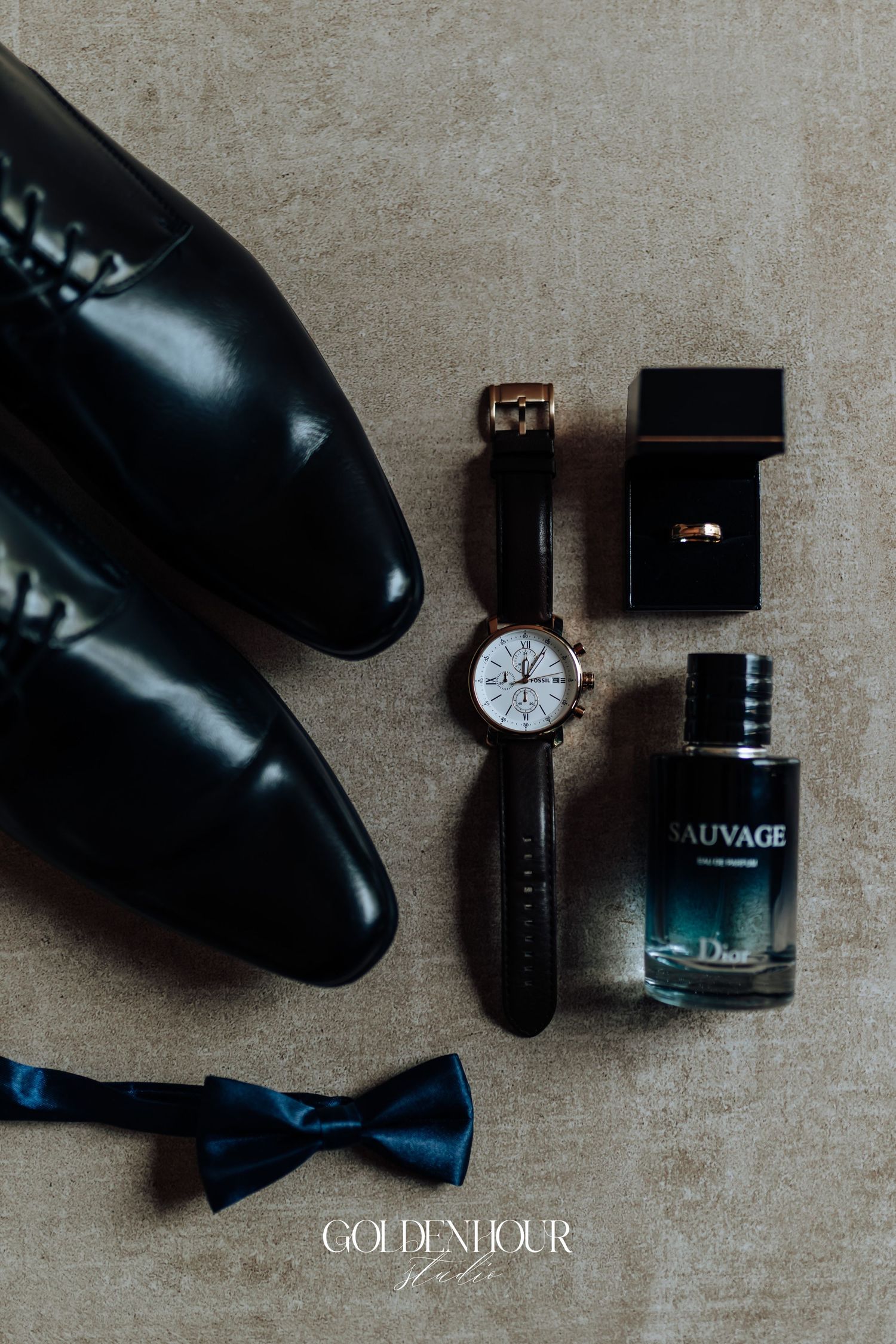 Wedding accessories including black dress shoes, bow tie, watch and cologne arranged on beige surface.