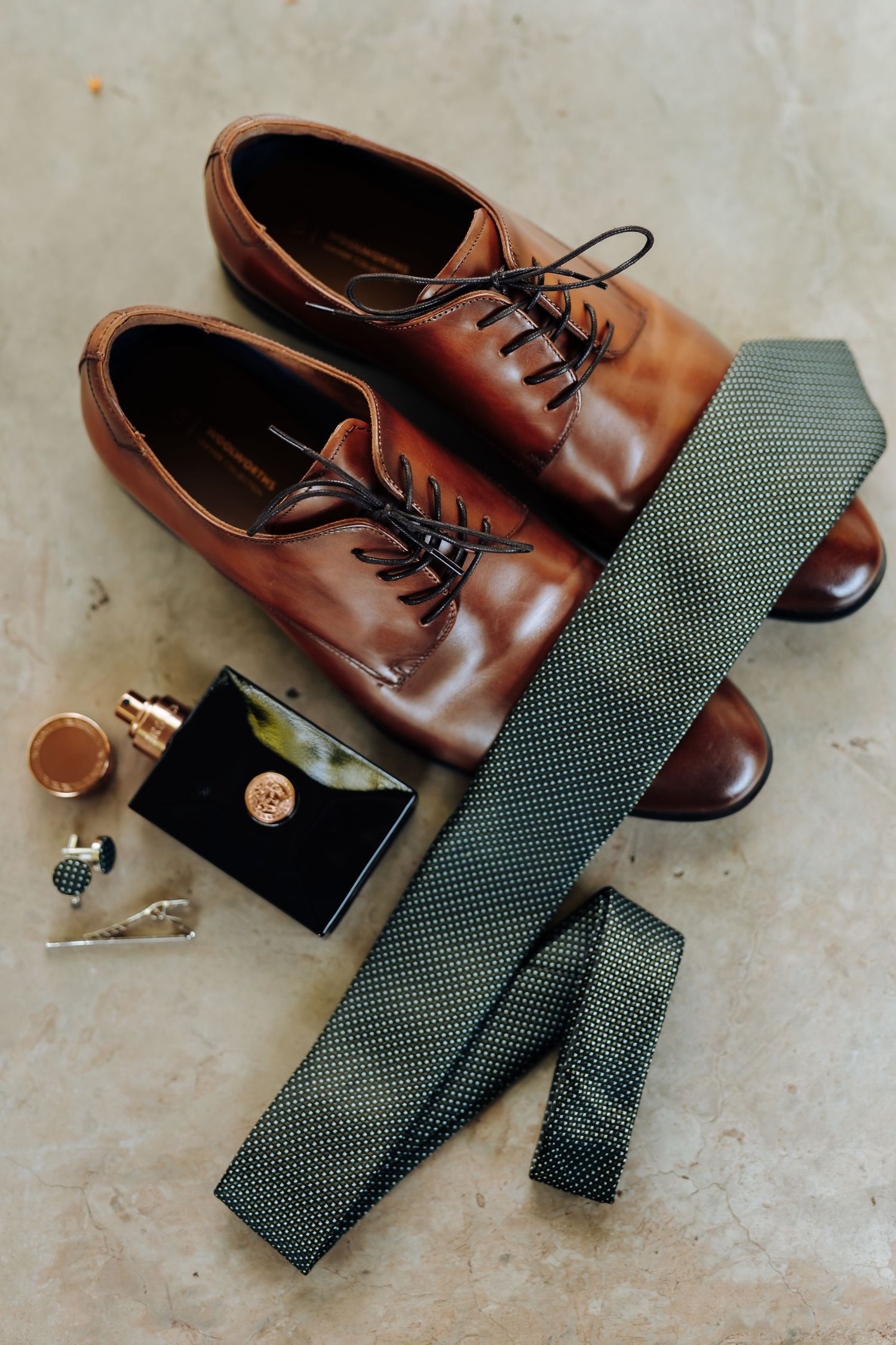 Brown leather dress shoes paired with a dark green patterned tie and accessories on a beige textured surface.