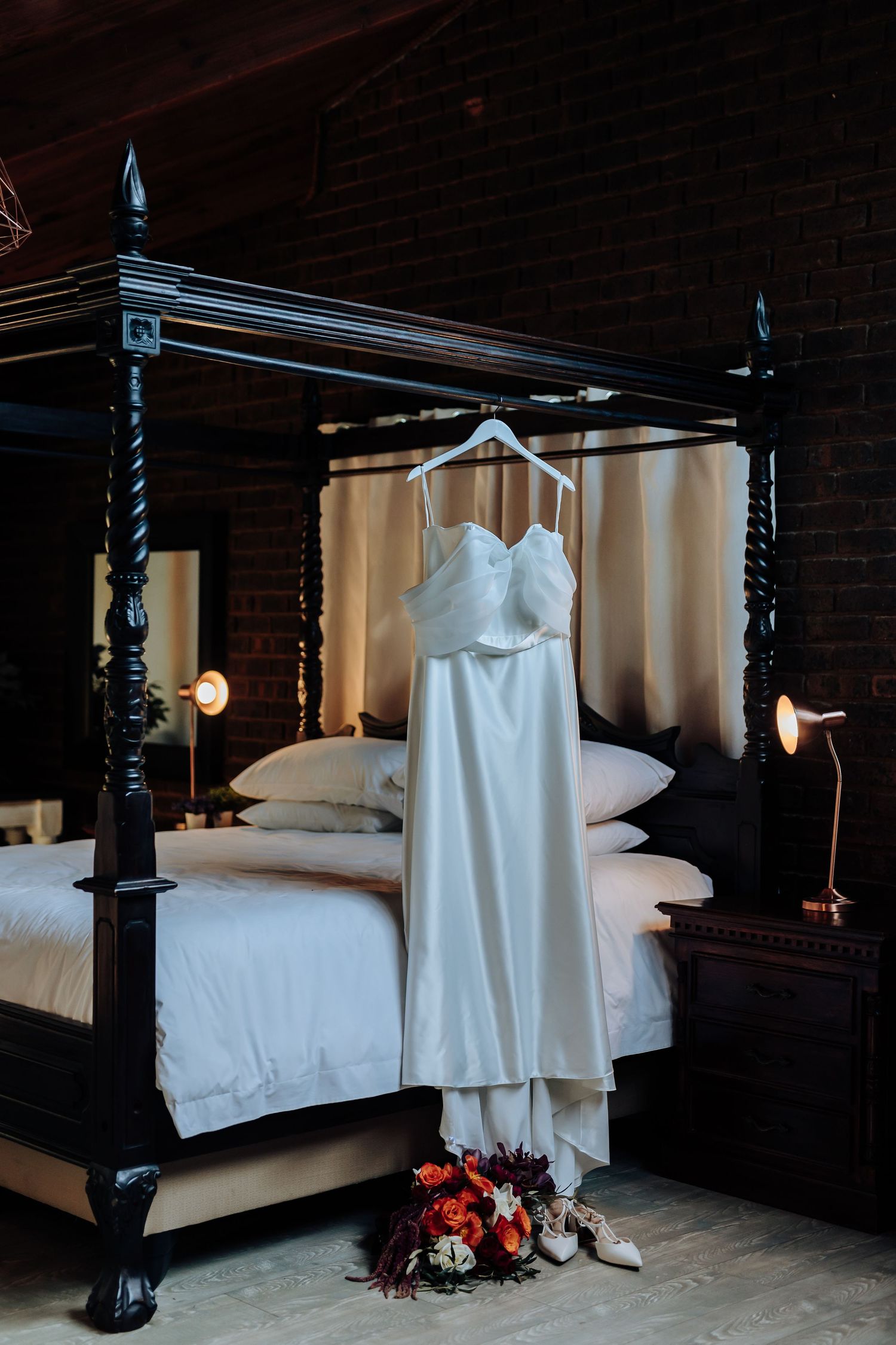 A white wedding dress hangs elegantly against dark wood walls and white bedding in a rustic bedroom setting.