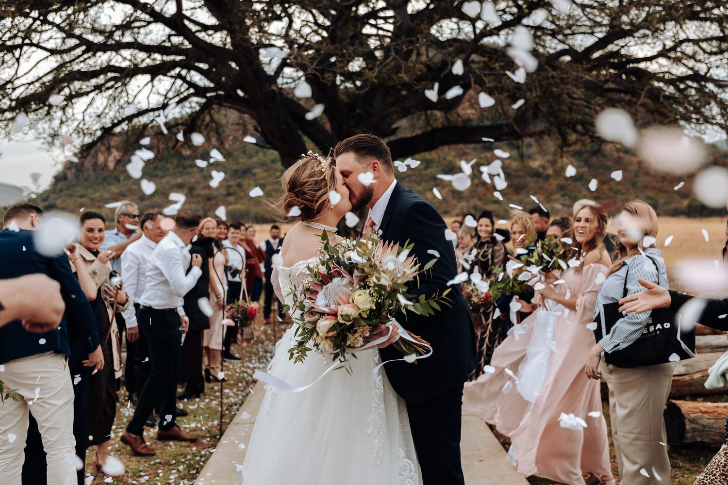 Newlywed couple shares intimate moment as wedding guests shower them with white confetti under a large tree.