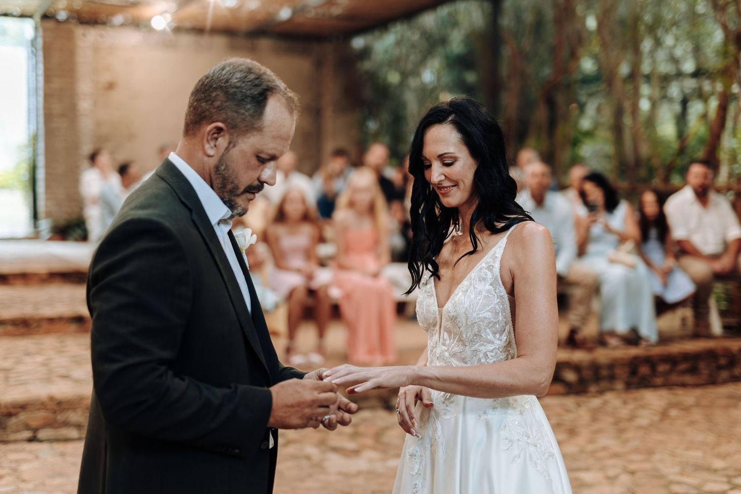 Bride and groom exchanging rings during outdoor wedding ceremony with guests seated in background.