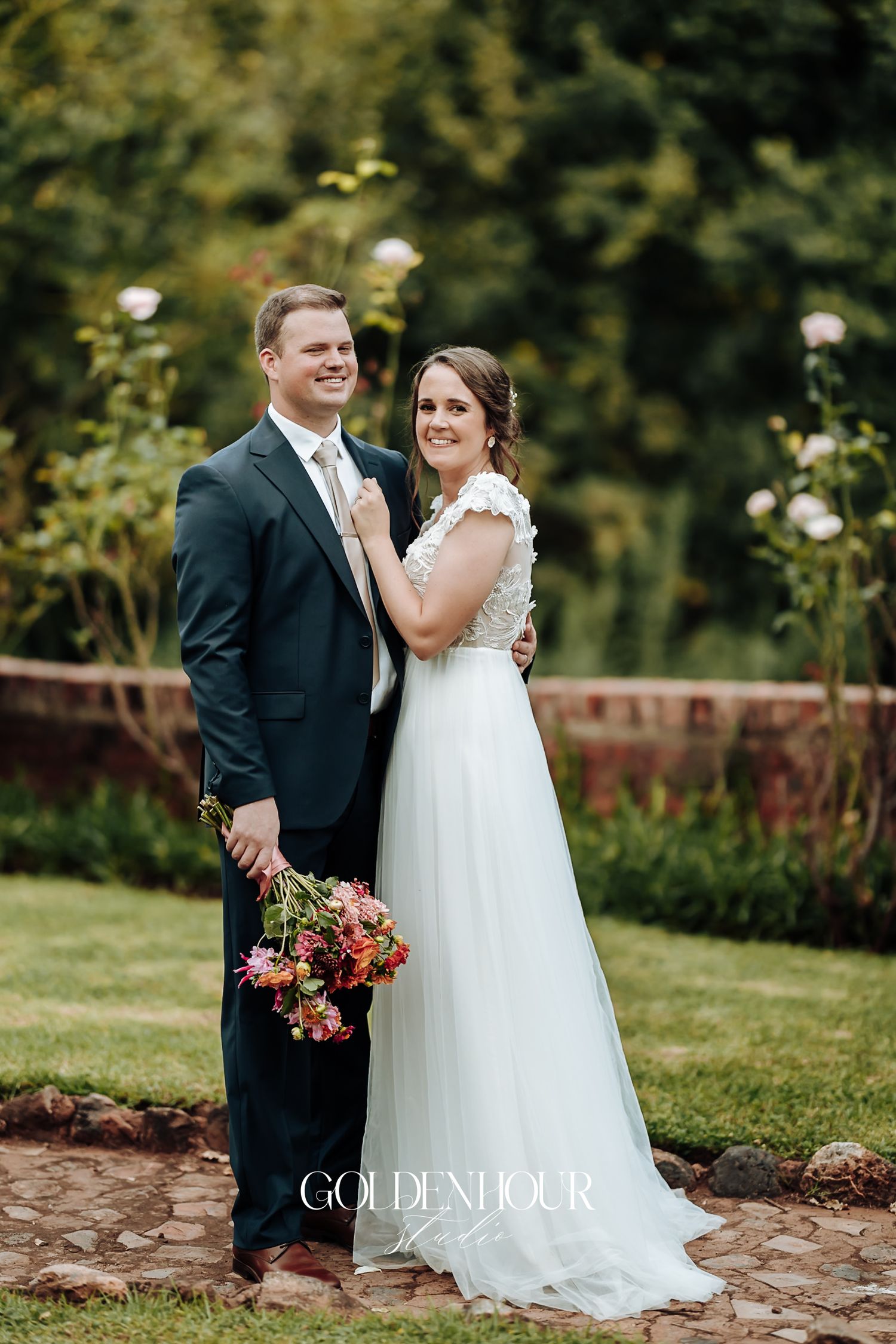 Bride and groom posing in rose garden, bride in lace gown holding colorful bridal bouquet.