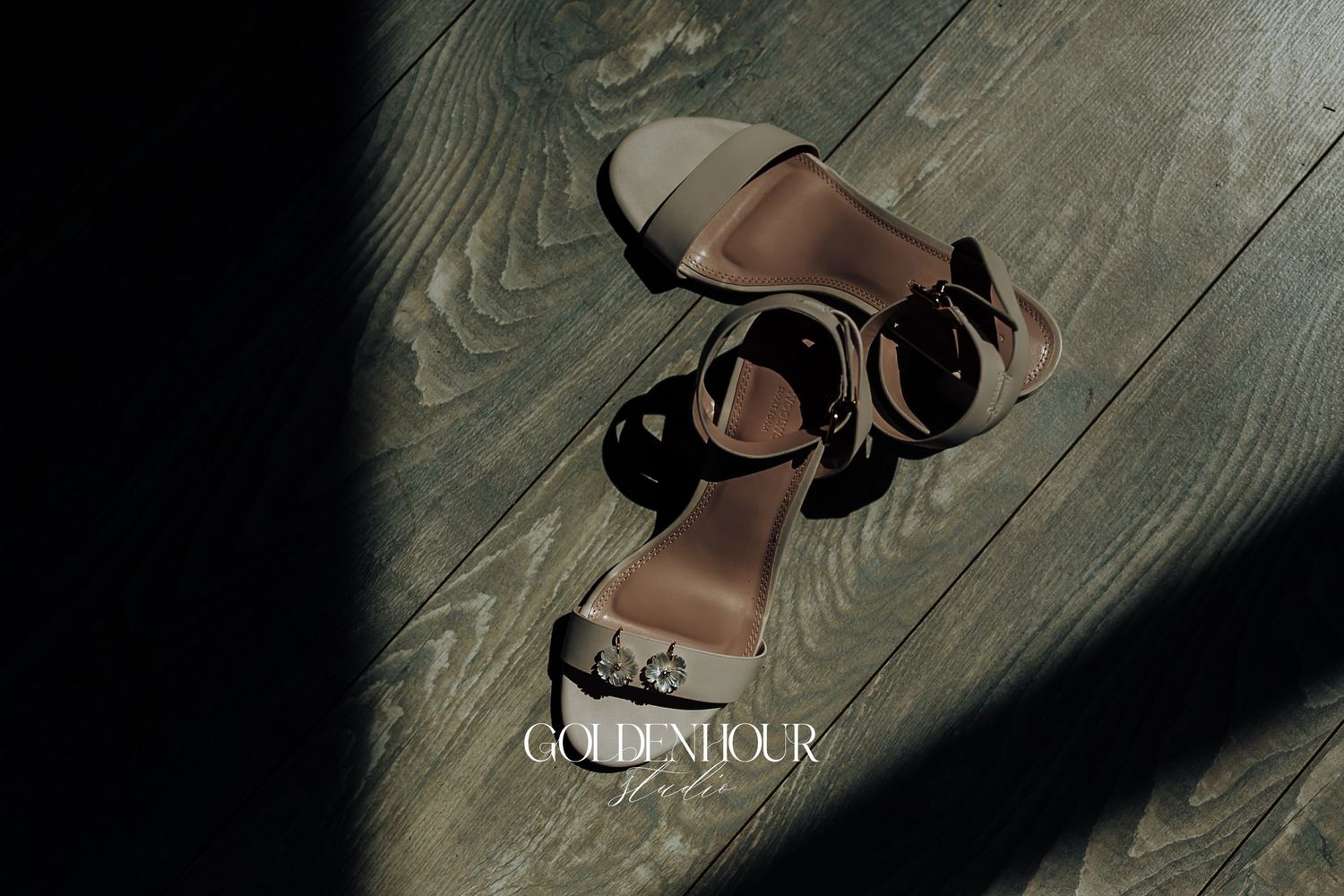 Strappy nude bridal heels resting on dark hardwood floor in dramatic lighting.