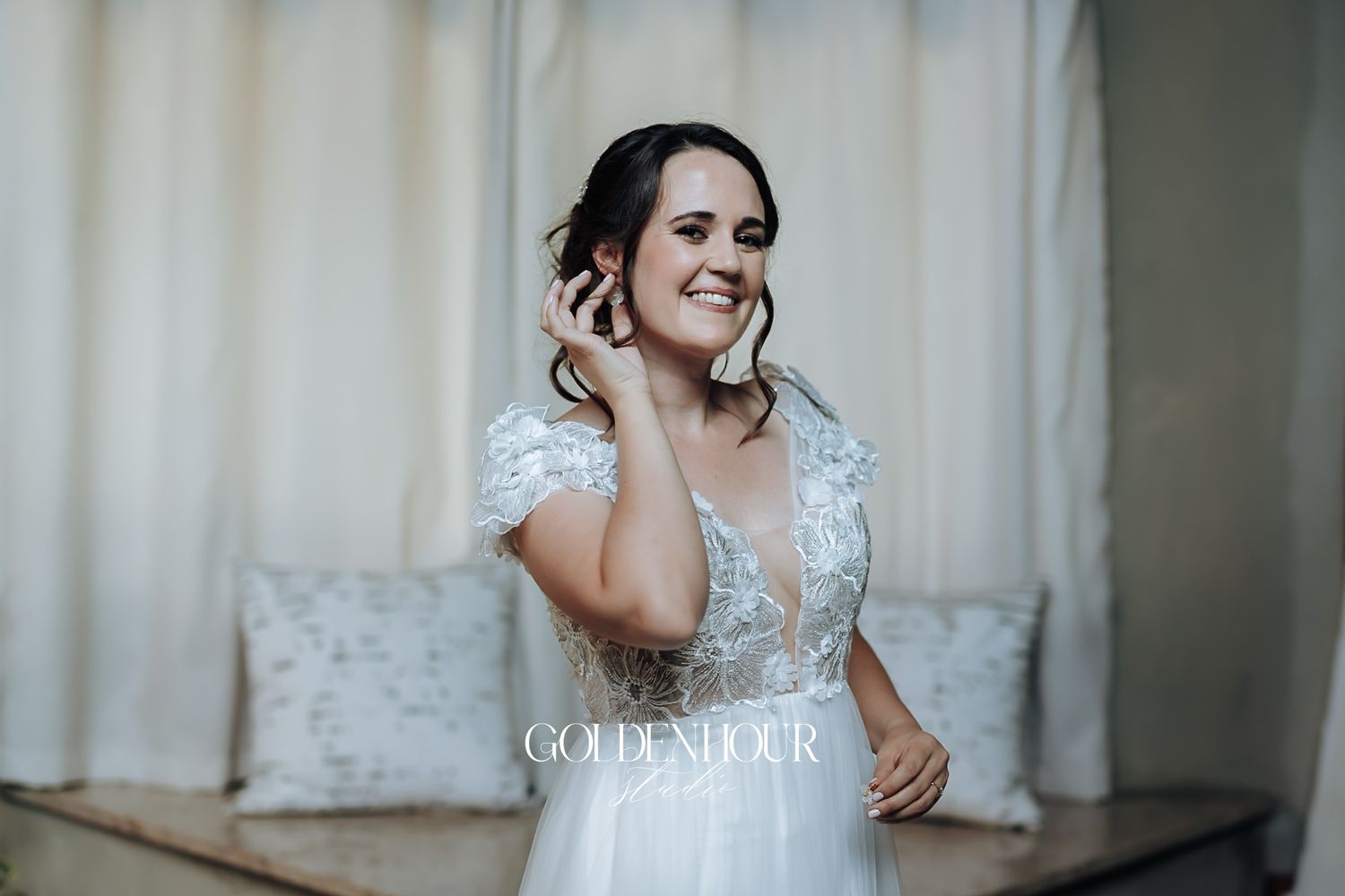 Bride in white lace wedding gown adjusting her earring while smiling beautifully.