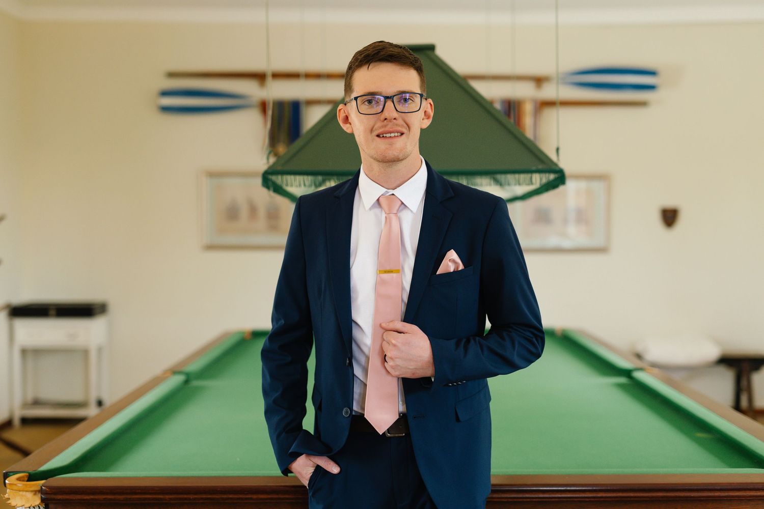 Professional wearing a navy suit and pink tie poses next to a pool table in an elegant game room setting.