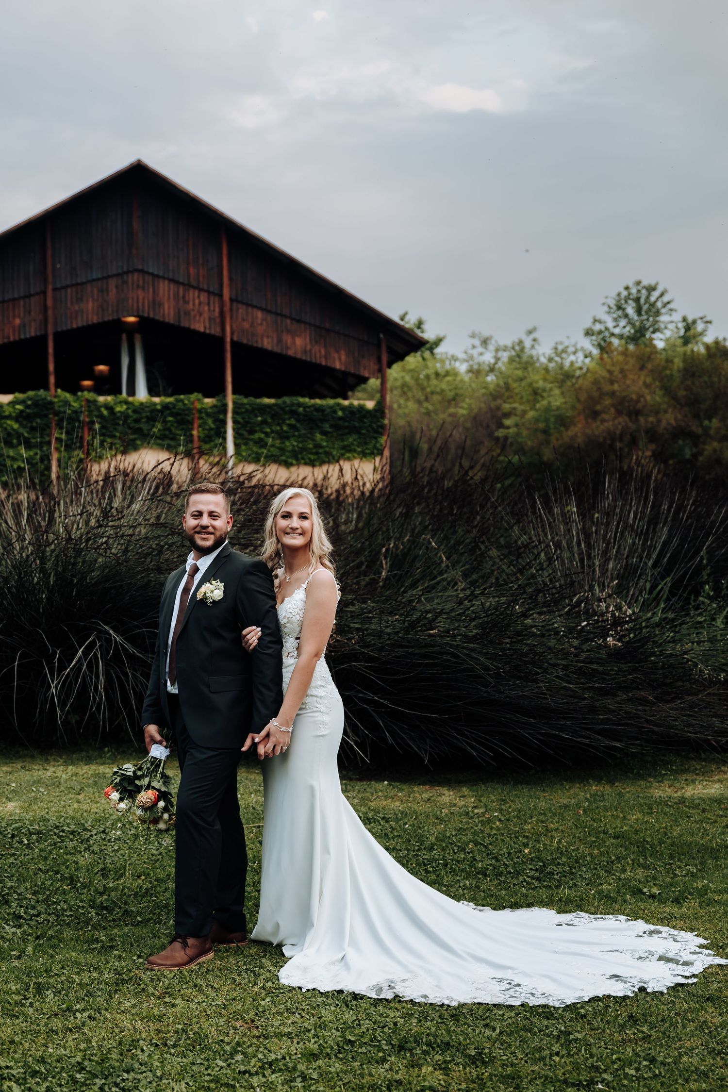 A couple in wedding attire poses in front of a rustic barn with tall grass and greenery in the foreground.