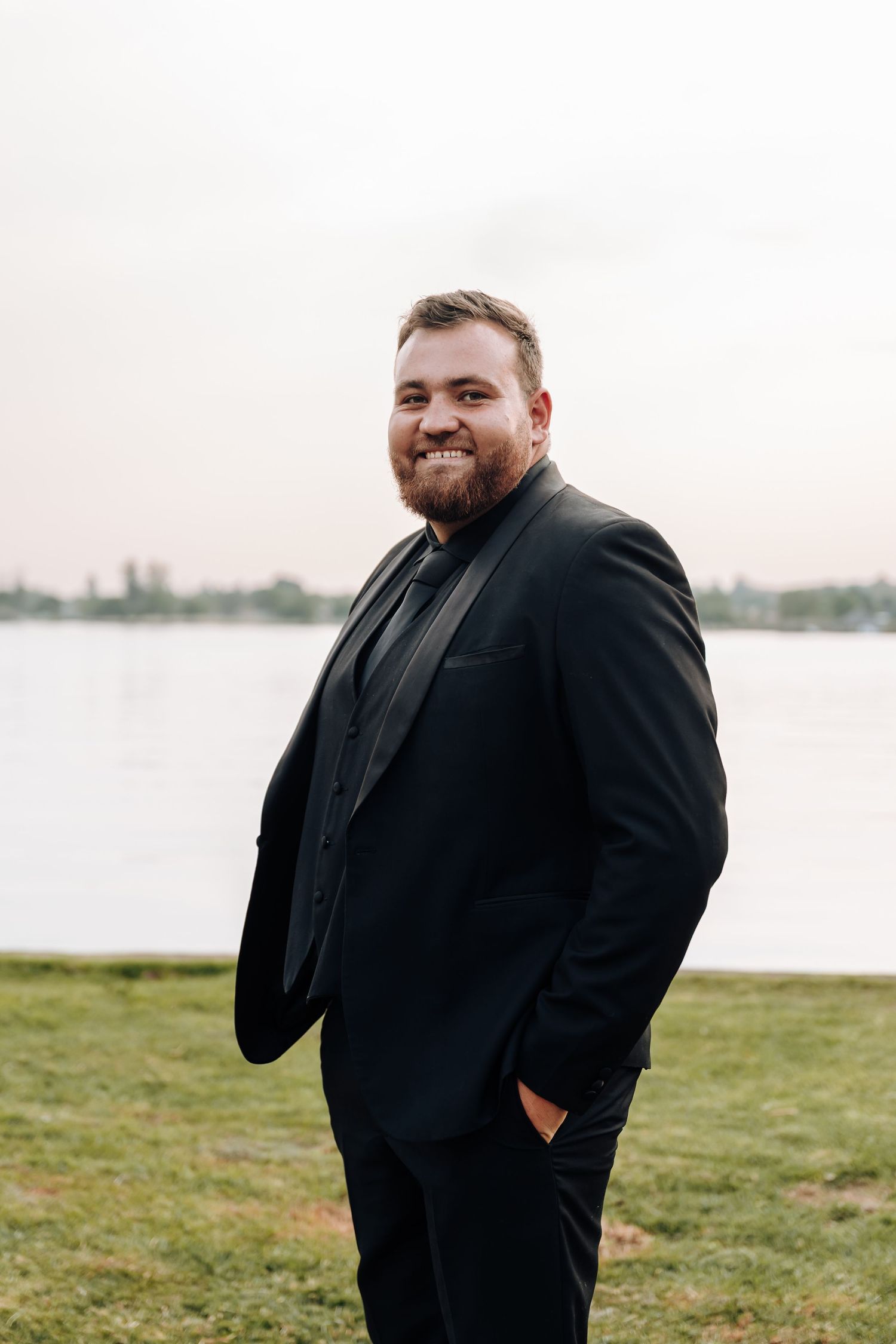 A person in a black suit stands outdoors on grassy waterfront area with a natural smile and relaxed pose.
