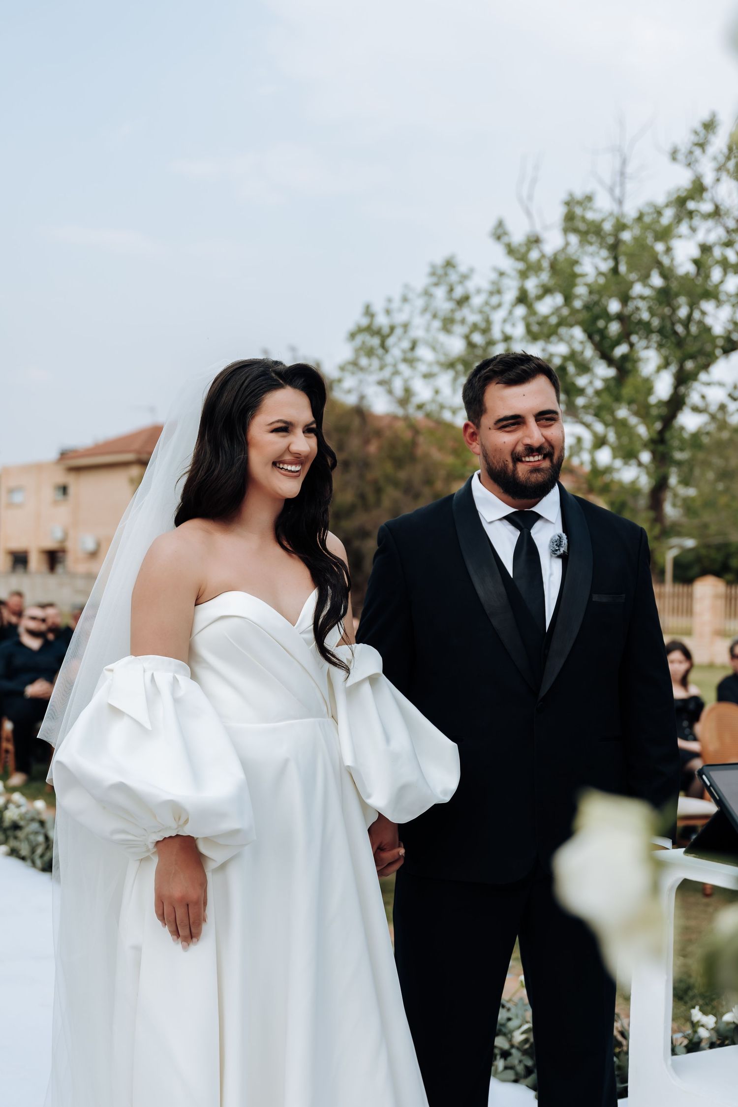 A bride in a white off-shoulder gown and groom in black suit share candid moments during their outdoor wedding.