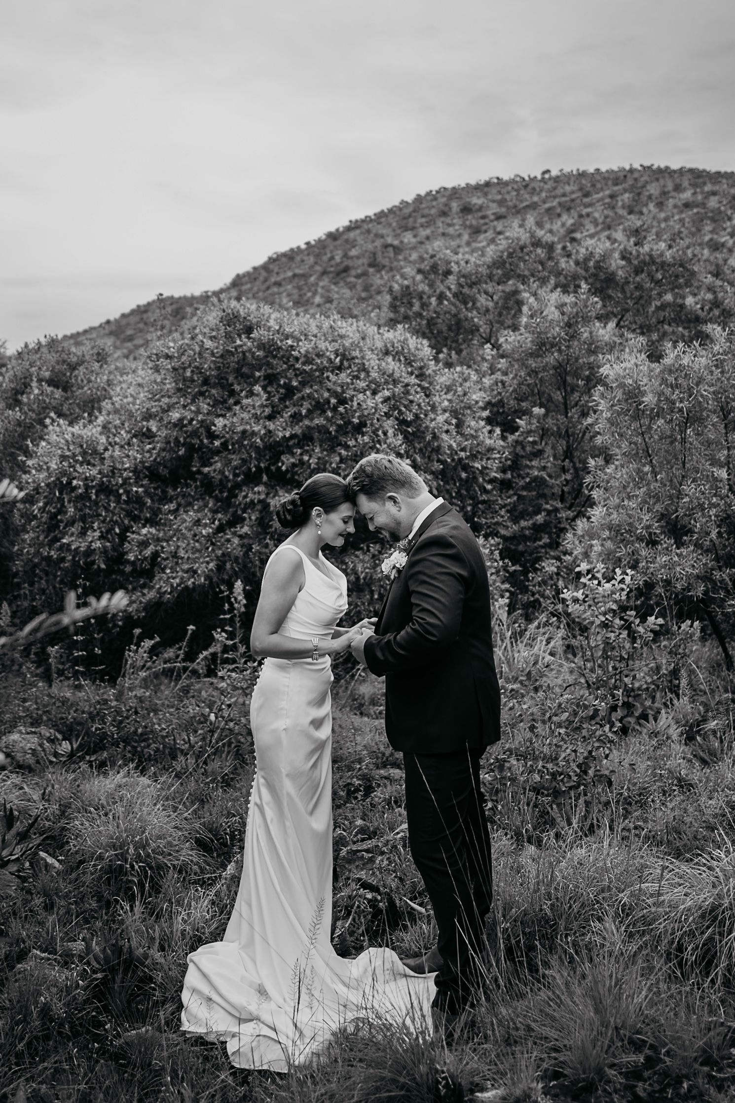A black and white wedding photo series shows a couple dancing together on a hillside surrounded by trees and foliage.