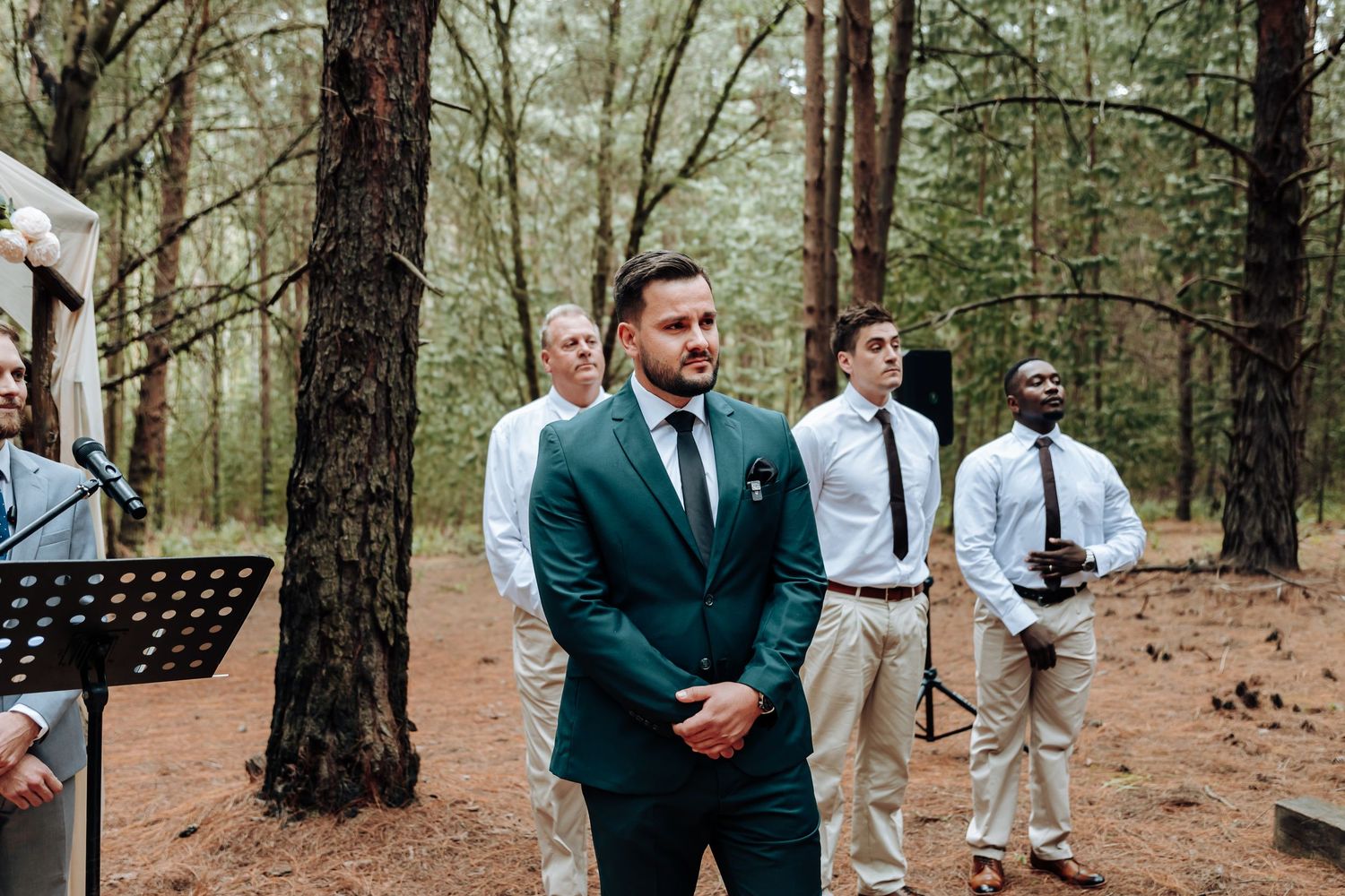 A groom in an emerald green suit stands during an outdoor forest wedding ceremony with guests in light attire behind him.