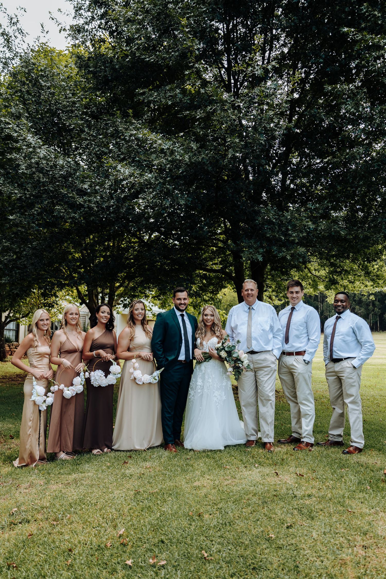 Wedding party in earth tones poses on grassy hill with bridesmaids in brown dresses and groomsmen in light suits.