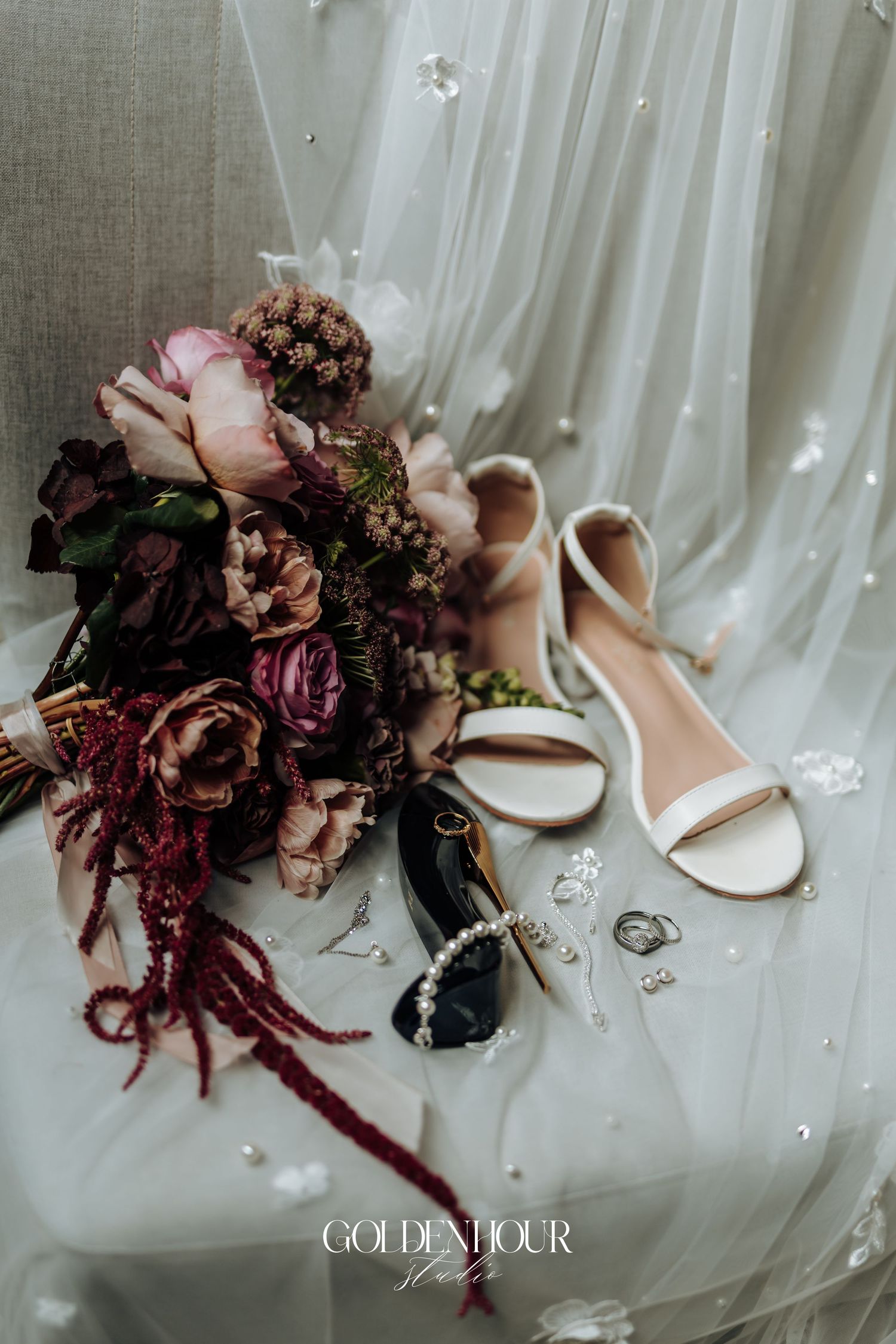 Bridal flat lay featuring a burgundy and pink bouquet, white heels, pearl jewelry, and rings on a white veil.