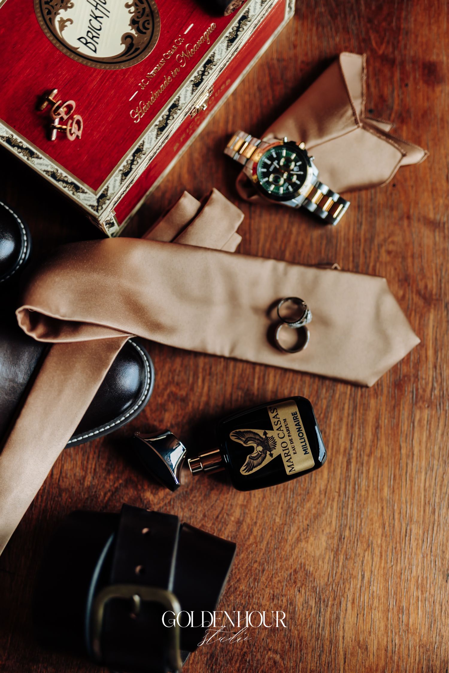 Groom's wedding flat lay featuring a tan tie, Rolex watch, rings, whiskey bottle, cufflinks, and cigar box on wood.