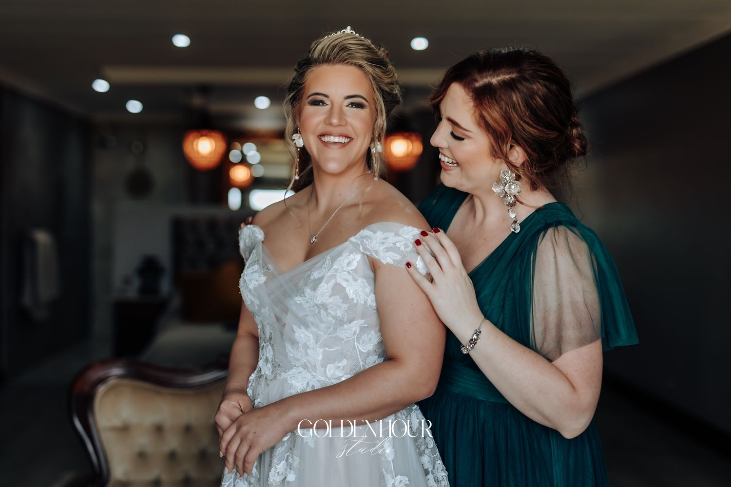 Bride in a lace wedding gown being helped by a bridesmaid in an emerald green dress indoors.
