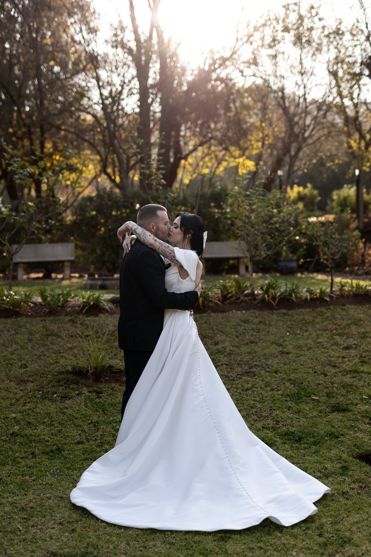 A couple embraces in a romantic dance at sunset in a garden setting with a flowing white wedding gown trailing behind them.