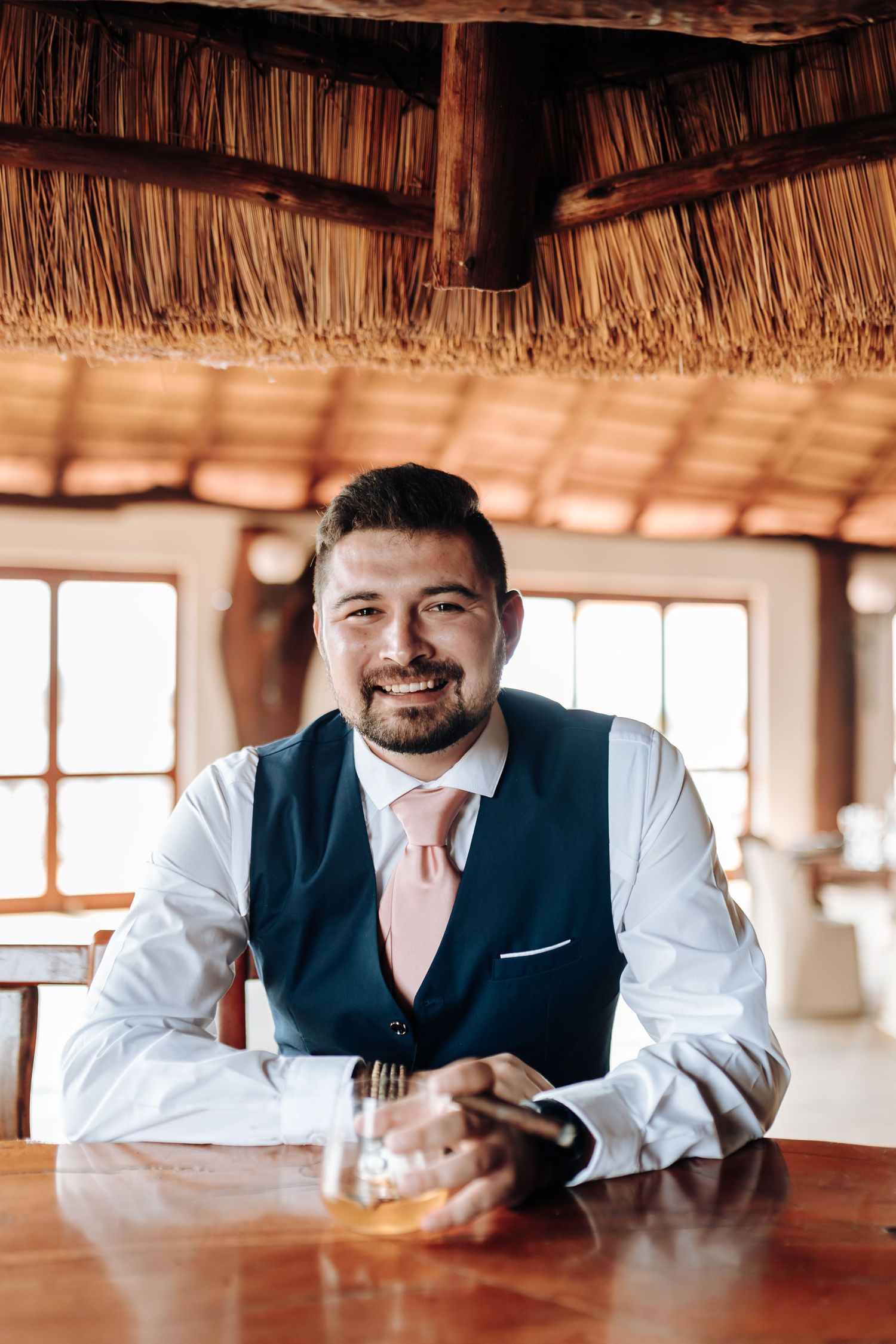A person in business attire sits smiling at a wooden table inside a tropical thatched-roof restaurant.