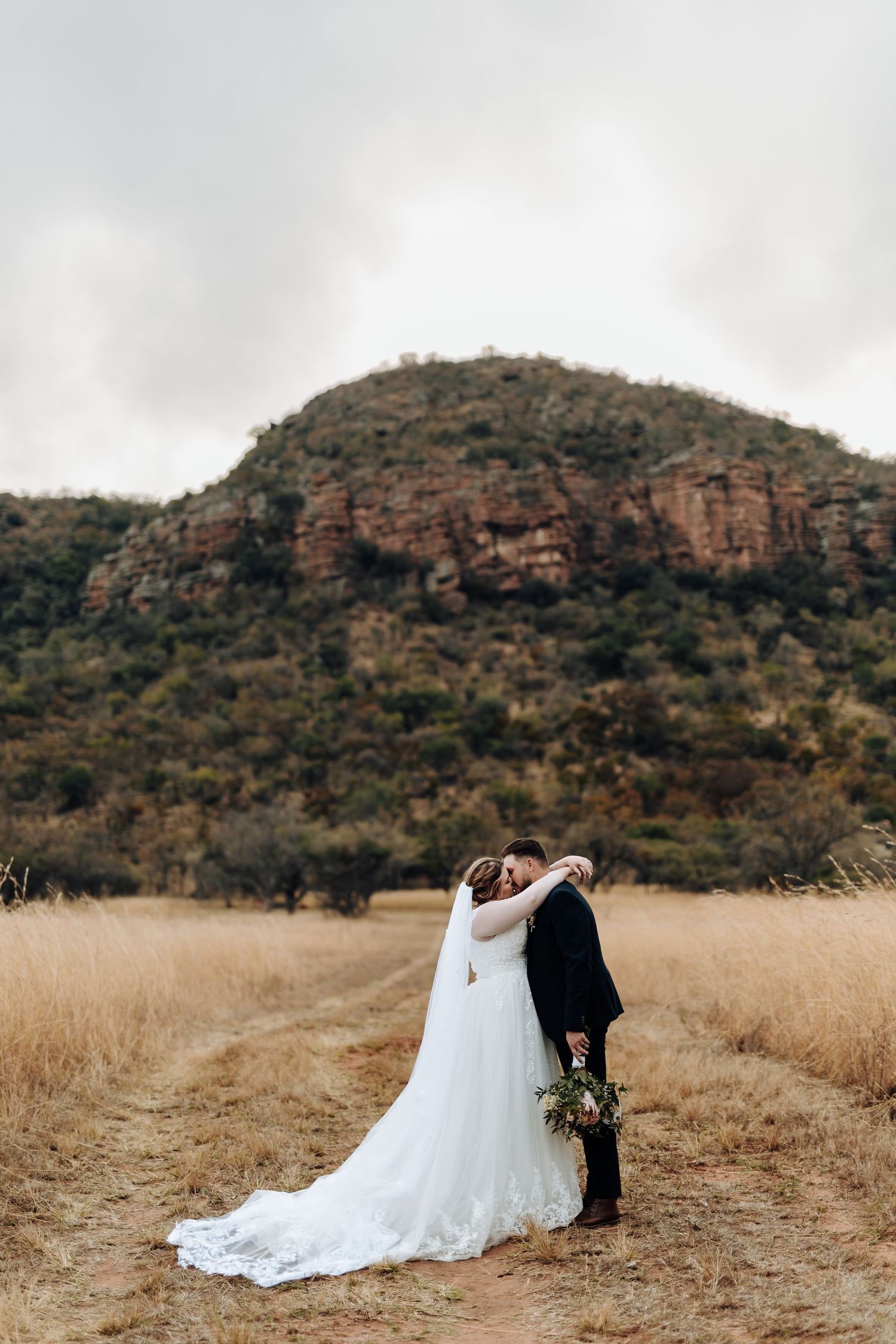 A romantic moment captured in a golden field with rocky mountains in the background, showcasing a flowing white wedding dress.