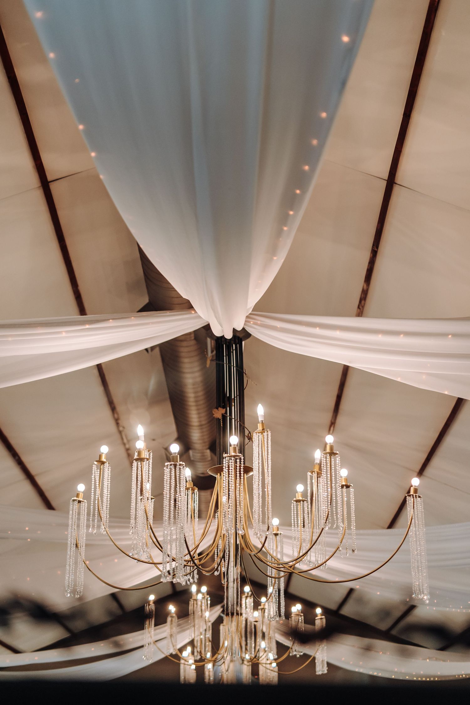 Elegant crystal chandelier hangs from white draped fabric ceiling in wedding reception venue with warm lighting.