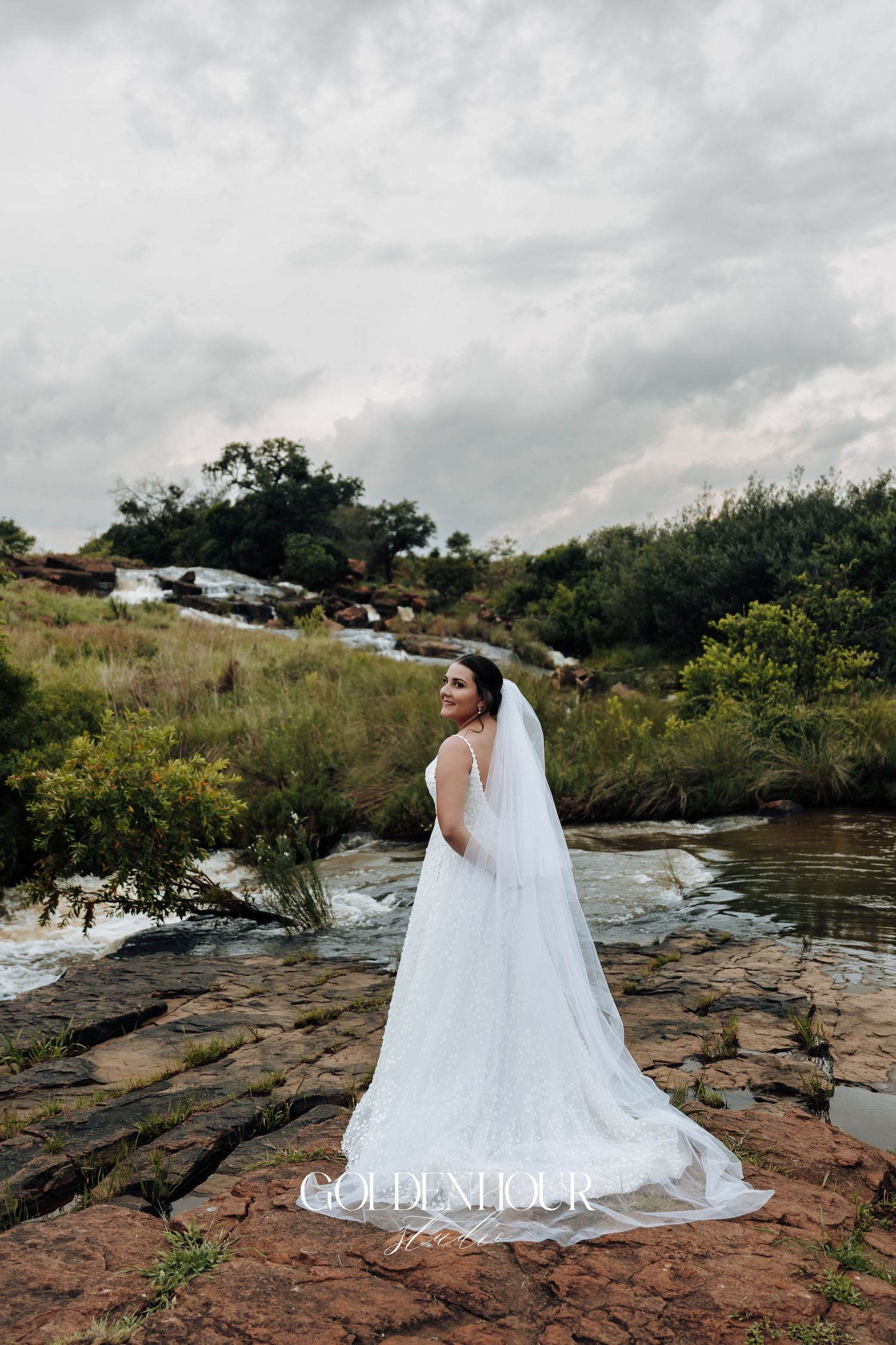Bride in flowing white dress poses on rocky terrain near water.