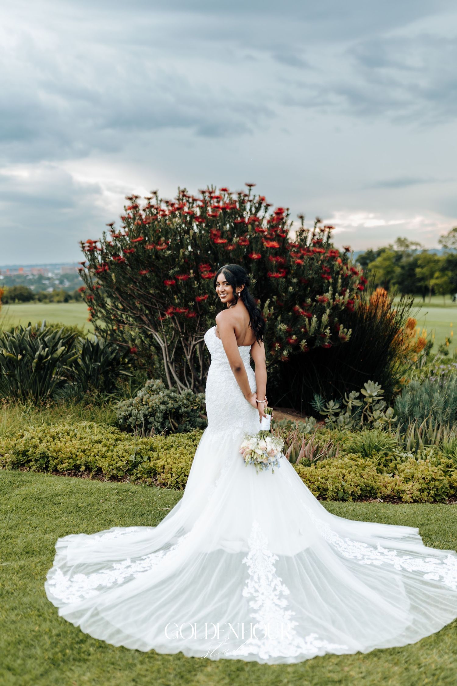 A stunning white mermaid wedding dress with long train photographed against vibrant red flowering bushes.