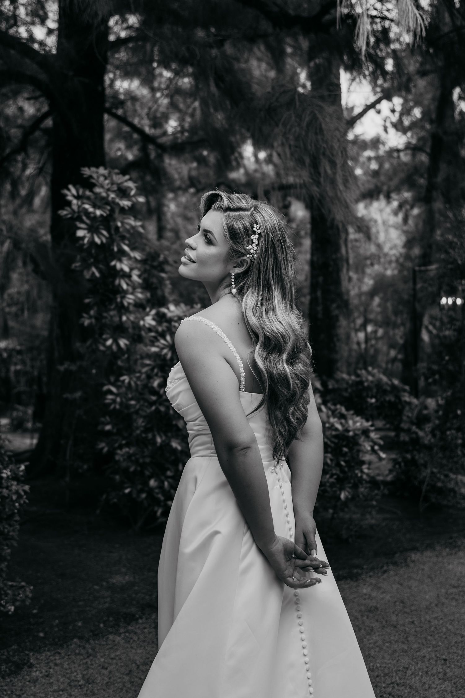 A bride in a white wedding dress stands in a forest setting with dramatic black and white lighting.