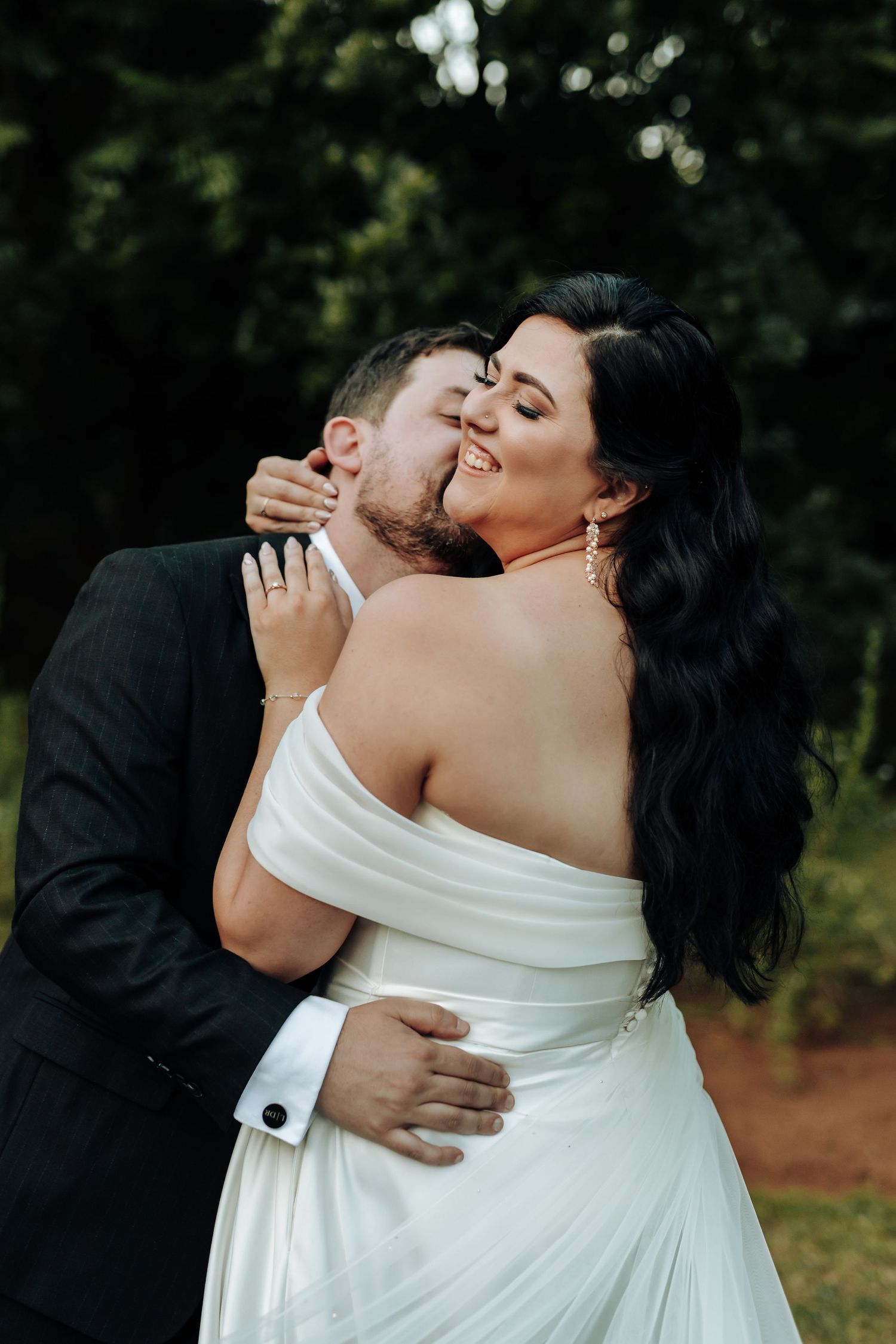 Romantic wedding couple embraces outdoors, with bride in white off-shoulder dress against natural backdrop.