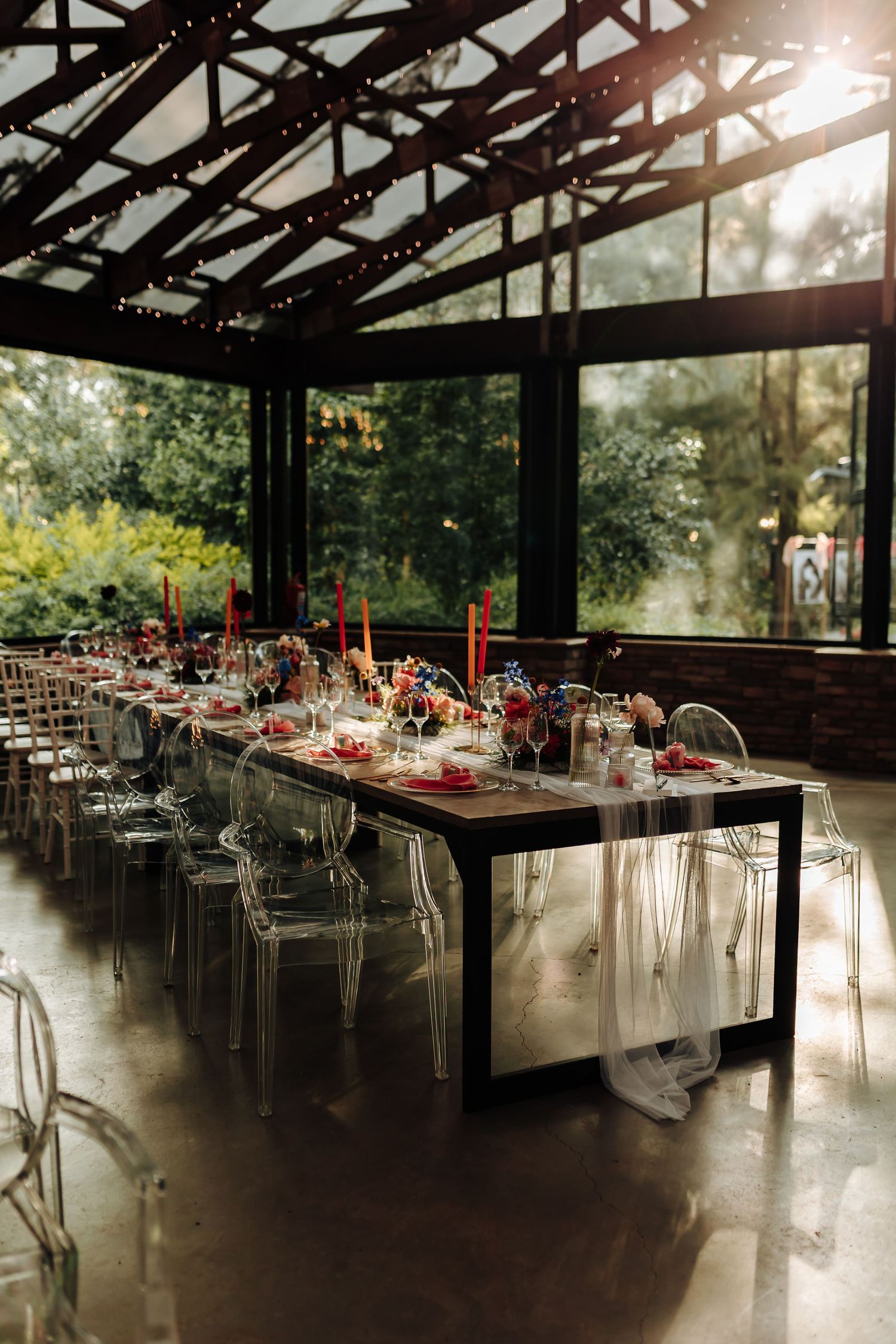 Long reception tables set with red floral centerpieces and ghost chairs in a rustic venue with natural lighting.