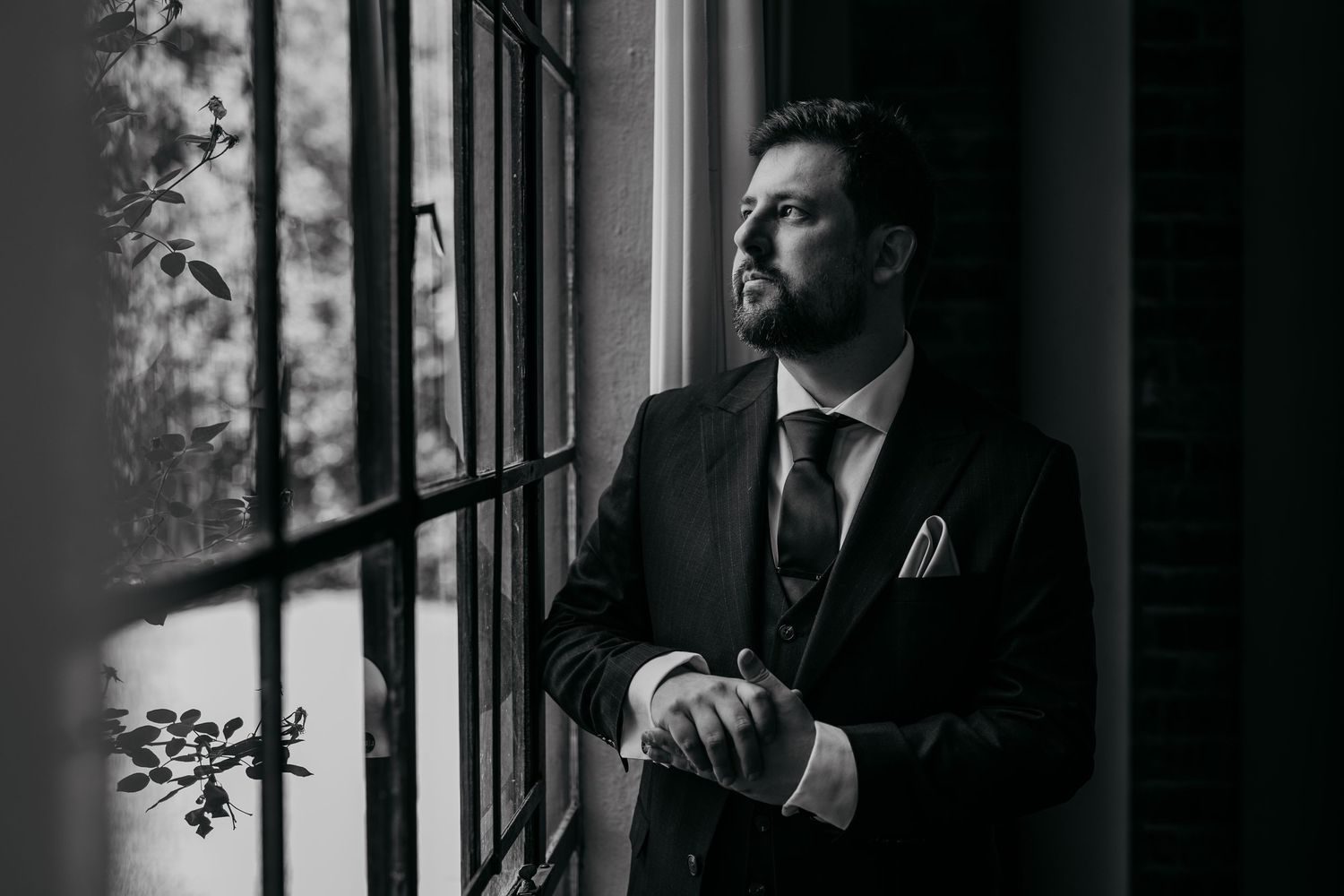 Dramatic black and white portrait of groom getting ready by a window in moody lighting.