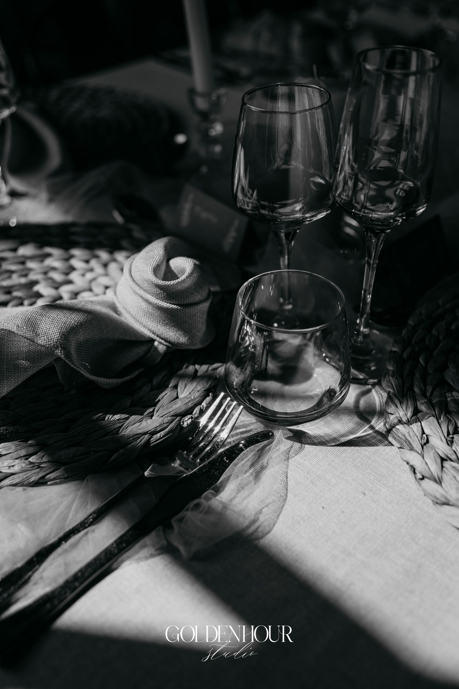 Black and white still life of elegant wine glasses and place settings on a table with dramatic shadows.