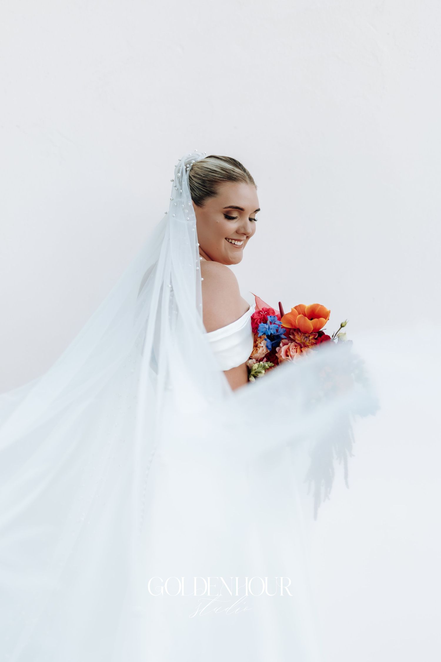 A bride in a flowing white veil holds a vibrant autumn-colored bouquet against a light background.