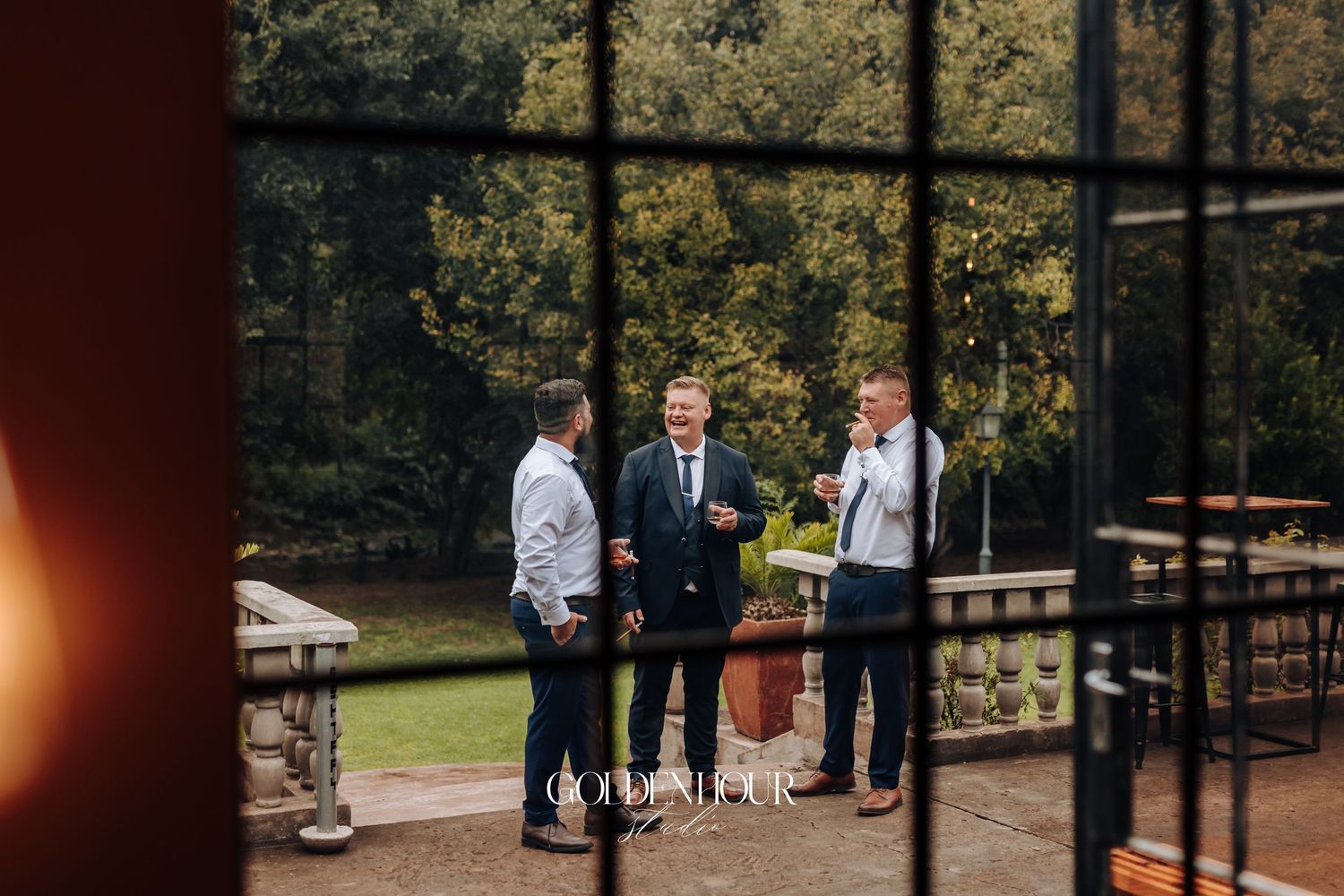 Groomsmen in suits laughing together in glass-walled venue garden before wedding ceremony.