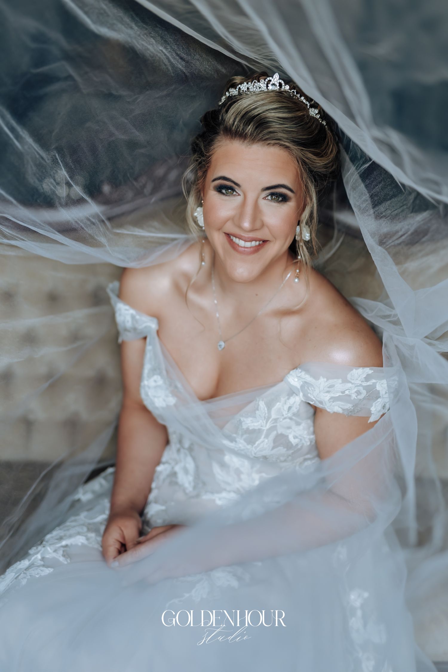 Radiant bride in off-shoulder lace gown with tiara smiling under flowing white veil in bridal portrait.